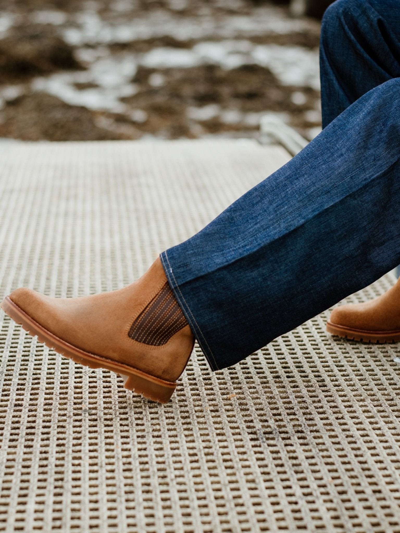 A person wearing blue jeans and Campbells of Beauly Leather Chelsea Boots with a durable rubber sole sits on a textured beige mat outdoors, blurred rocks and snow in the background.