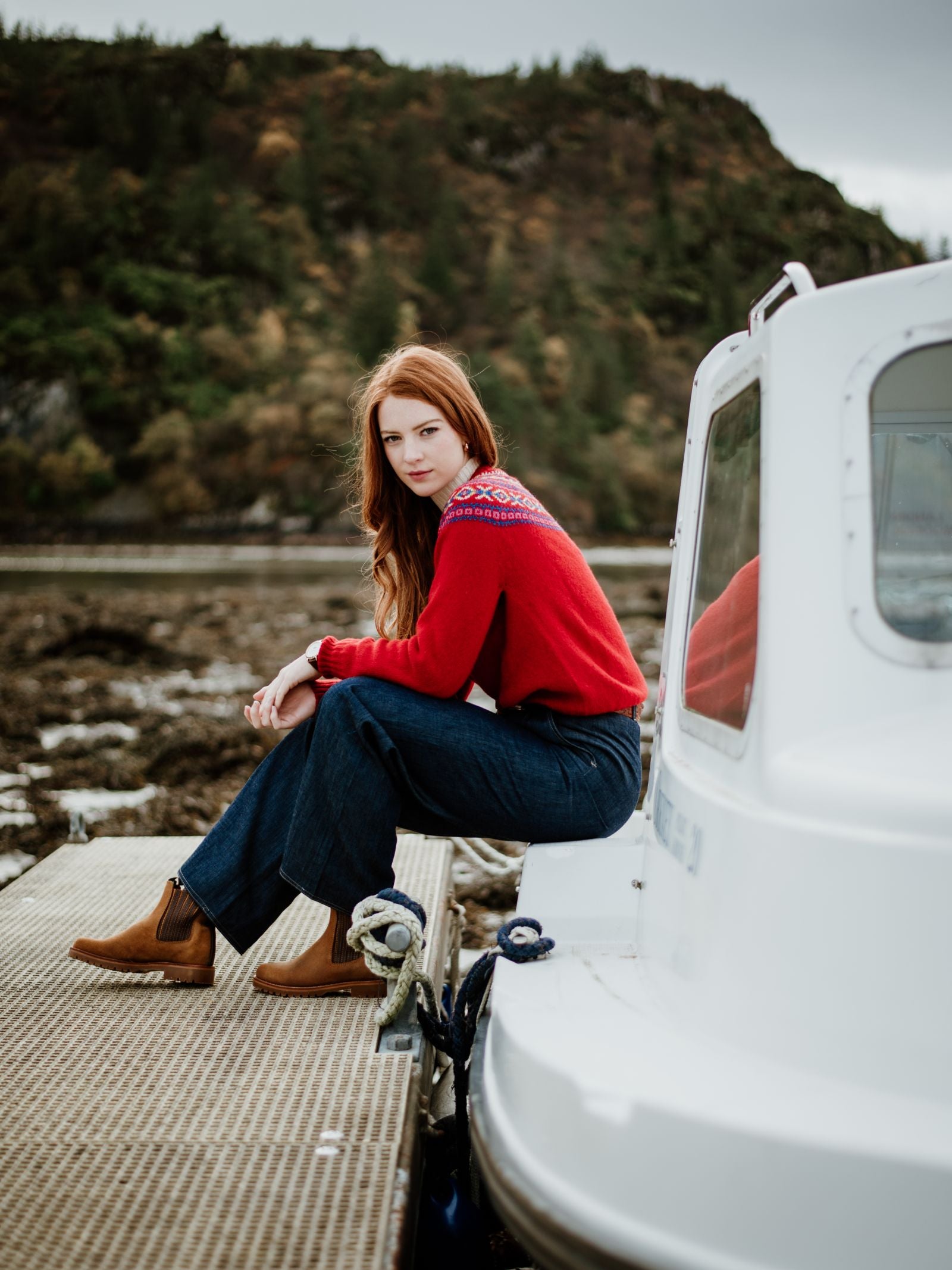 A young woman with long red hair sits on a dock by a white boat, wearing a red sweater, dark jeans, and Campbells of Beauly Leather Chelsea Boots with a durable rubber sole, against a forested hillside backdrop.