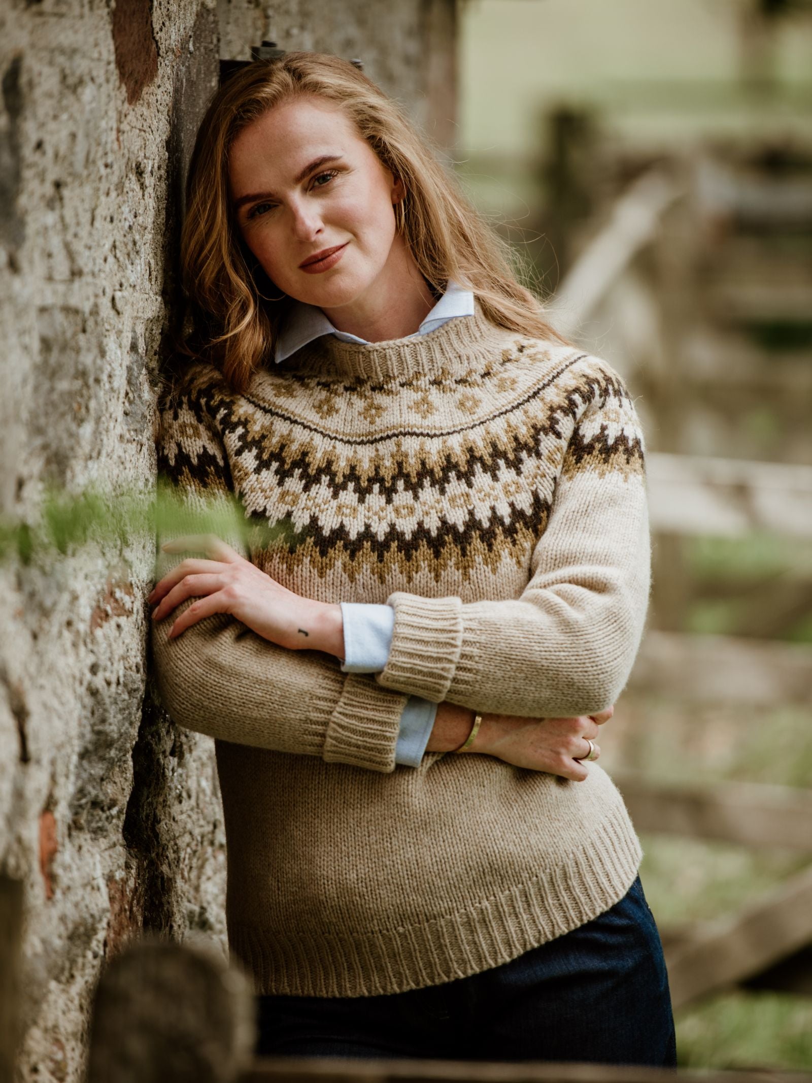 A woman with long, light brown hair leans against a stone wall outdoors, wearing the Campbells of Beauly Chunky Fairisle Jumper over a light blue collared shirt, arms crossed and looking relaxed.