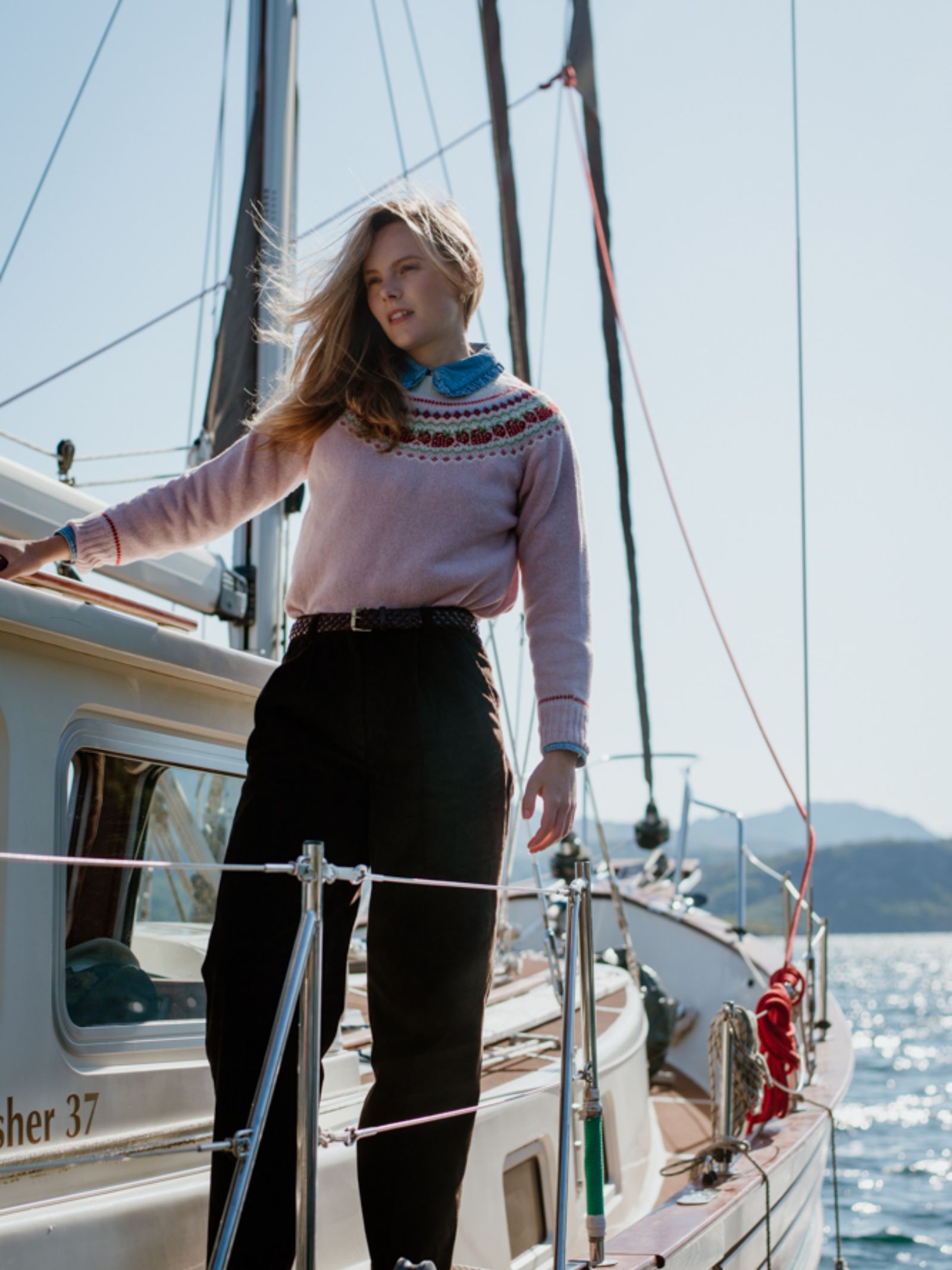 A woman with long blonde hair, wearing the Campbells of Beauly Geelong Lambswool Strawberry Yoke Jumper and black pants, stands on a sailboat deck on a sunny day, with water and distant hills in the background.