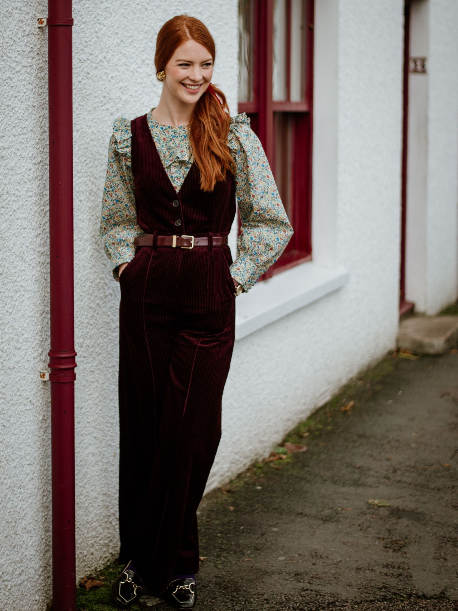A woman with long red hair smiles outdoors in a Campbells of Beauly Liberty Print Peplum Shirt, paired with a dark burgundy vest and pants, standing by a white wall with red window frames.
