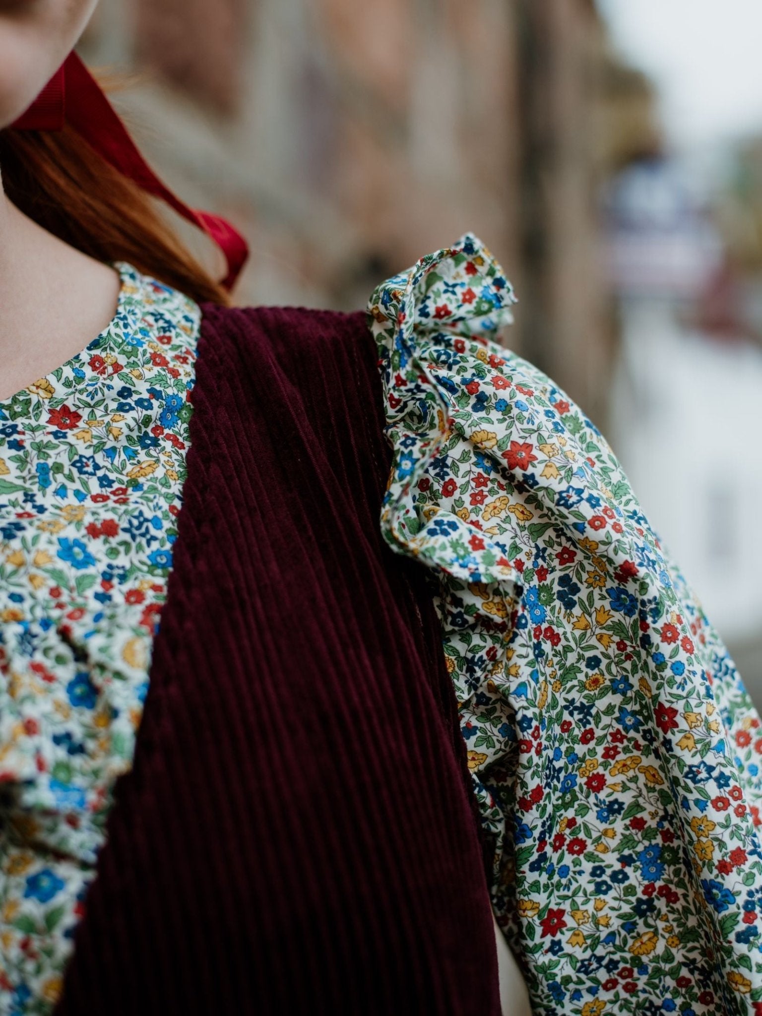 Close-up of a person in a Campbell’s of Beauly Liberty Print Peplum Shirt and deep burgundy corduroy vest, with ruffled sleeves visible. Only part of their head and shoulder appears against a blurred outdoor background.