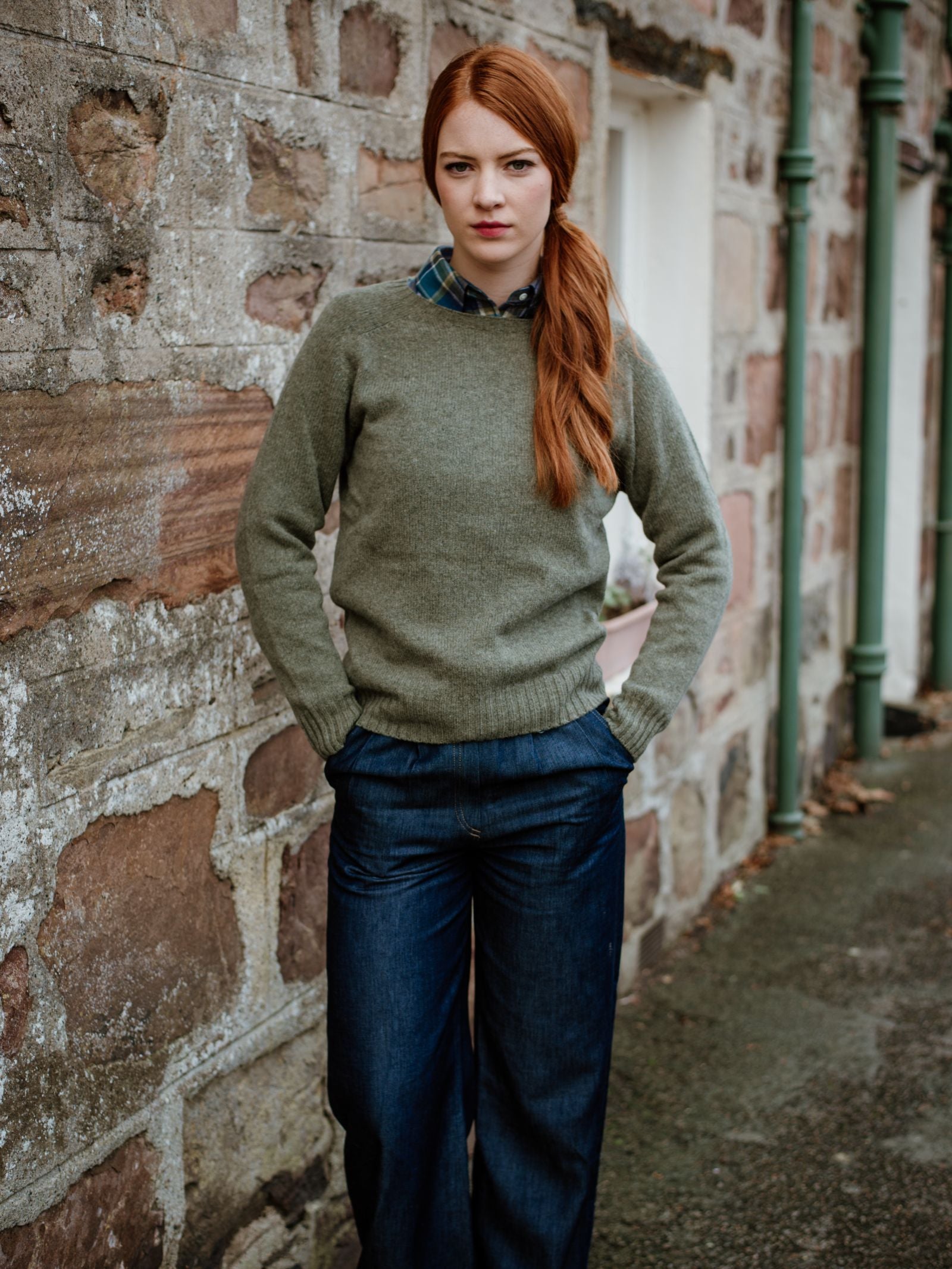 A woman with long red hair in a side ponytail stands by a stone wall in Scotland, wearing the Campbells of Beauly Geelong Lambswool Crew Neck Jumper over a plaid shirt and wide-leg jeans, hands in her pockets on a cloudy day.