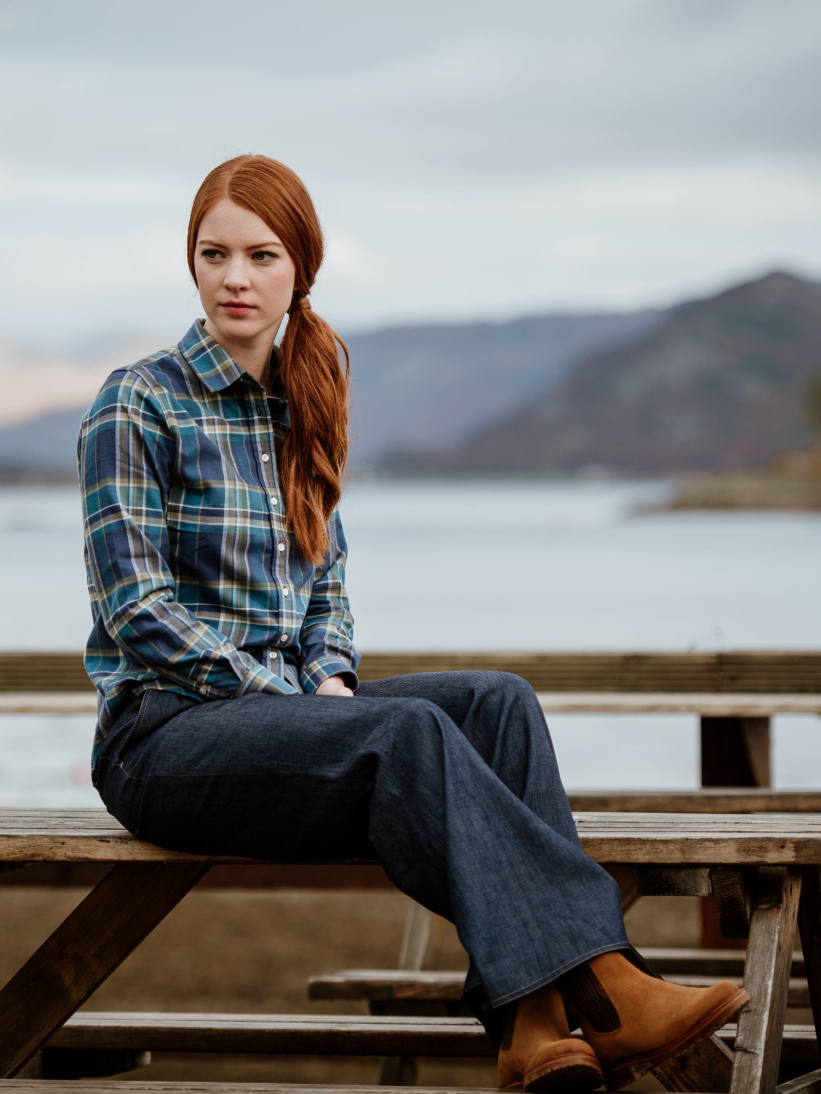 A woman with long red hair, in a plaid shirt, dark jeans, and Campbells of Beauly Leather Chelsea Boots with a durable rubber sole, sits on a wooden picnic table by a lake with mountains under cloudy skies.