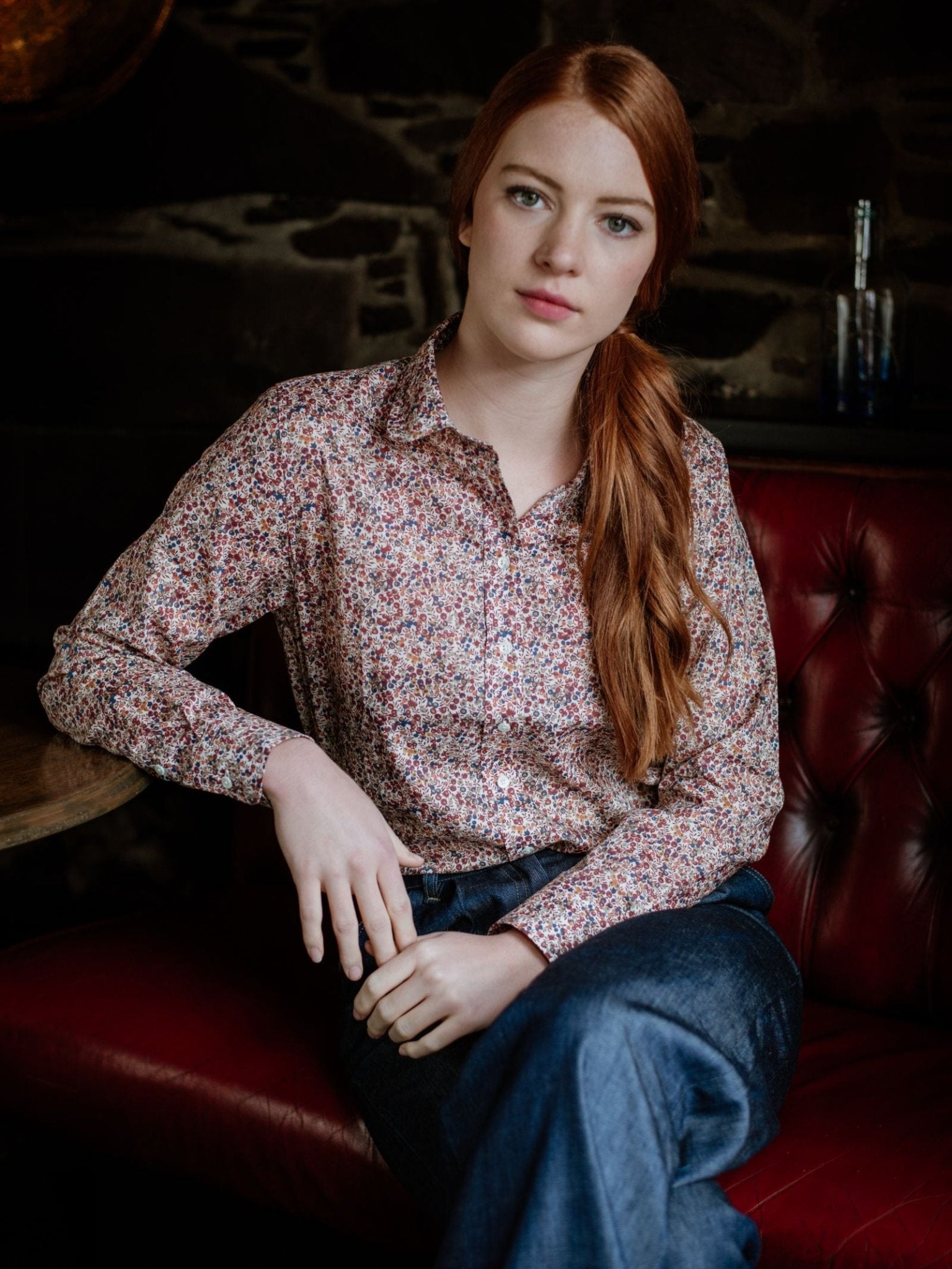 A woman with long red hair, dressed in a Campbells of Beauly Liberty Print Cotton Shirt and blue jeans, sits calmly on a red leather bench against a dark stone wall, gazing at the camera.