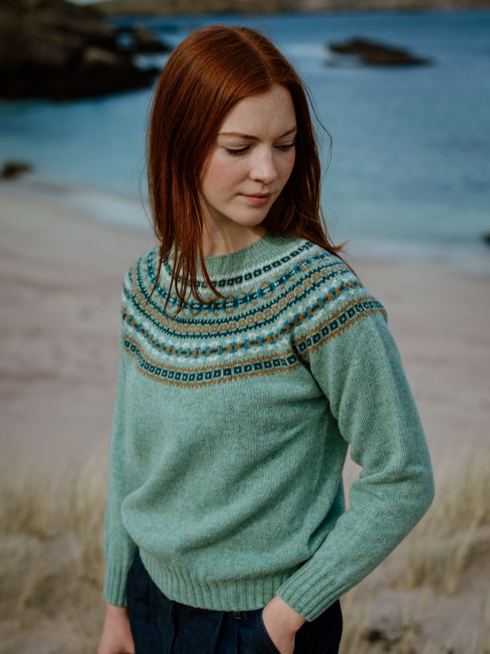 A young woman with long red hair stands on a beach, wearing a light green Campbells of Beauly Fairisle Crew Jumper with a traditional pattern. She looks down with her hands in her pockets, while the sea and rocks are visible behind her.