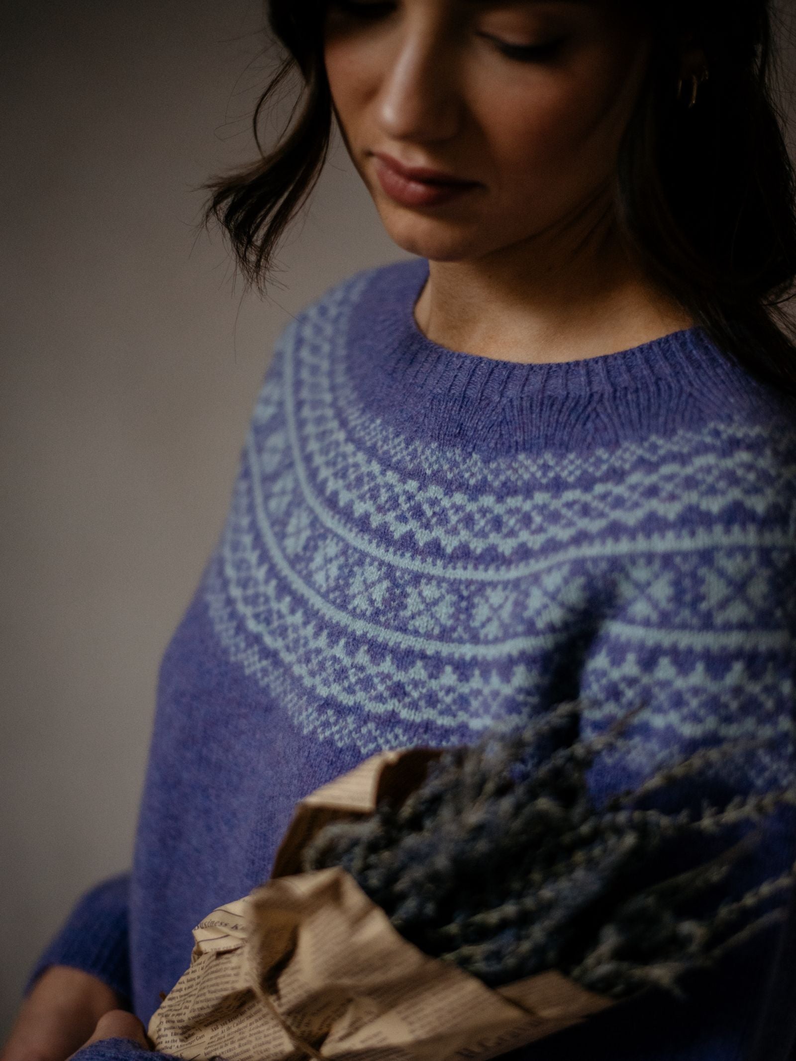 A woman wearing a blue Campbells of Beauly Two-Colour Fairisle Yoke Jumper holds a bouquet of dried lavender wrapped in brown paper, softly lit with focus on her upper body and the lavender.