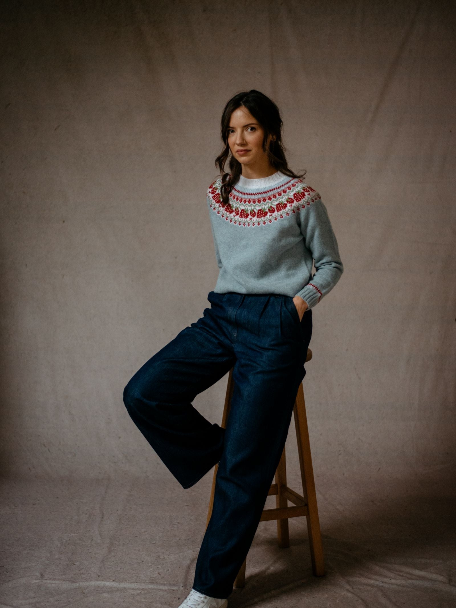 A woman with long brown hair sits on a wooden stool, wearing the Campbells of Beauly Geelong Lambswool Strawberry Yoke Jumper, wide-leg navy pants, and white sneakers against a plain beige background.
