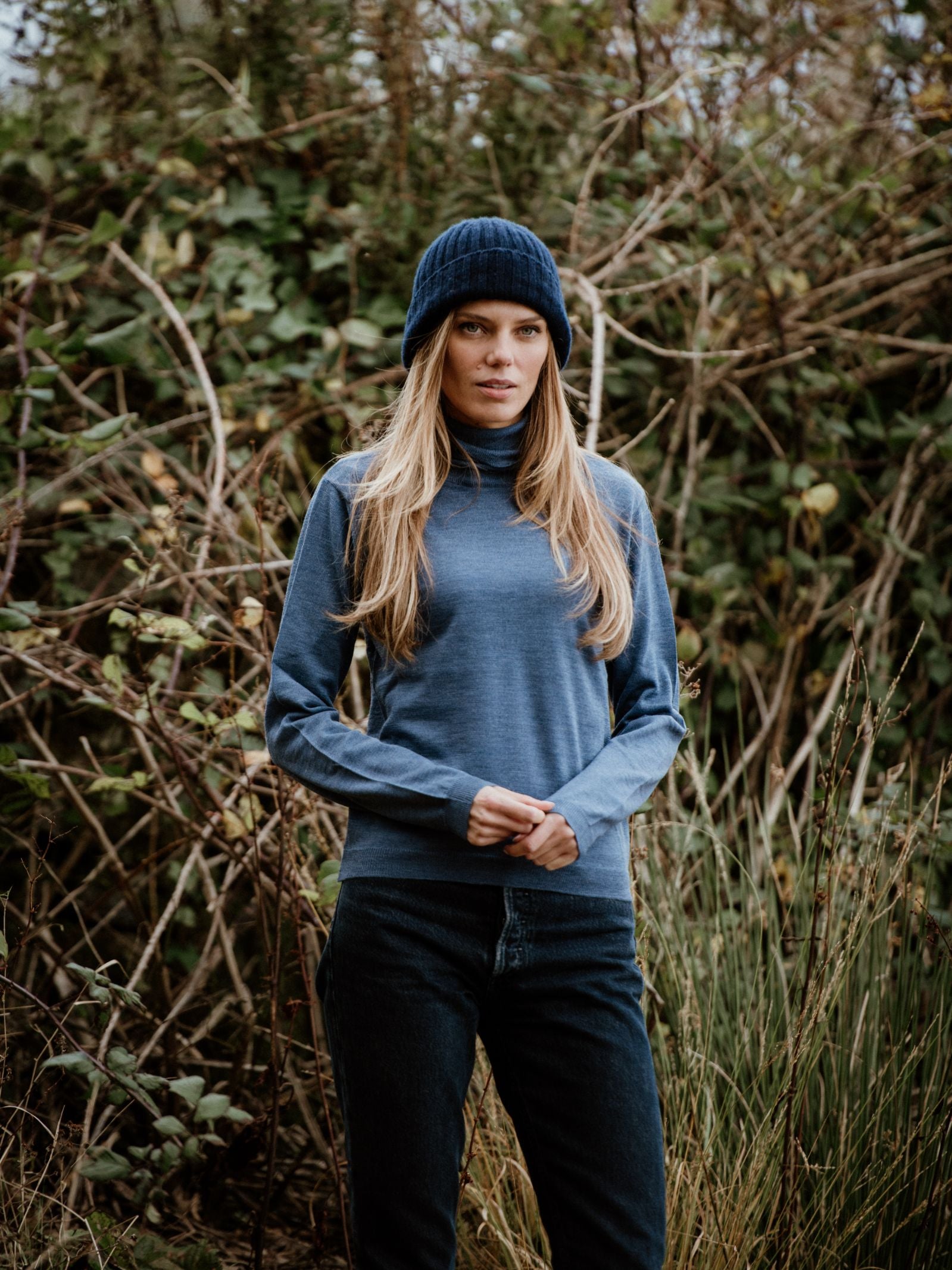 A woman with long blonde hair, wearing the Campbell's of Beauly Auld Stock Merino Poloneck and a navy knit beanie, stands outdoors in front of dense, leafy bushes.