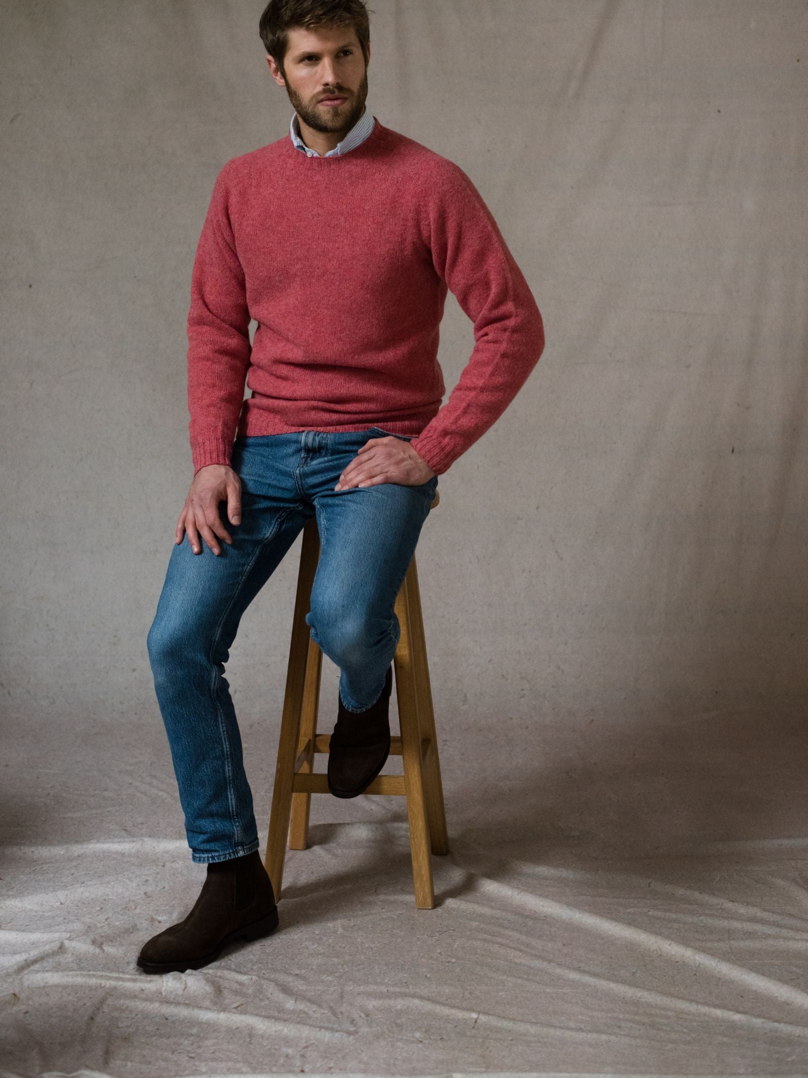 A bearded man sits on a wooden stool against a neutral backdrop, wearing the Campbells of Beauly Shetland Jumper in heritage-inspired colors over a collared shirt, blue jeans, and brown boots.