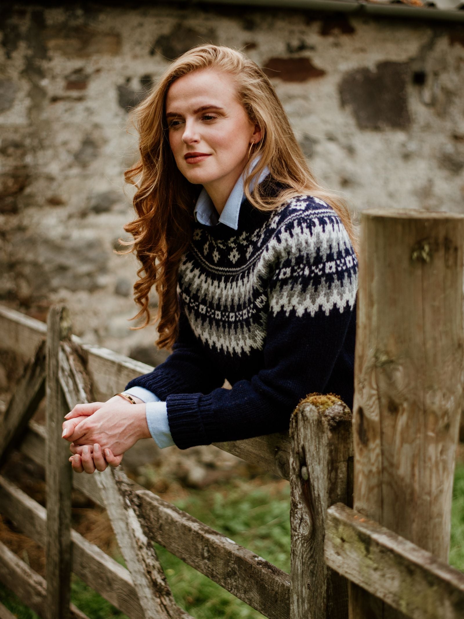 A woman with long red hair leans on a wooden fence, wearing the Campbells of Beauly Chunky Fairisle Jumper over a light blue shirt. Outdoors by a weathered stone wall, she looks thoughtful—her go-to winter staple for chilly days.
