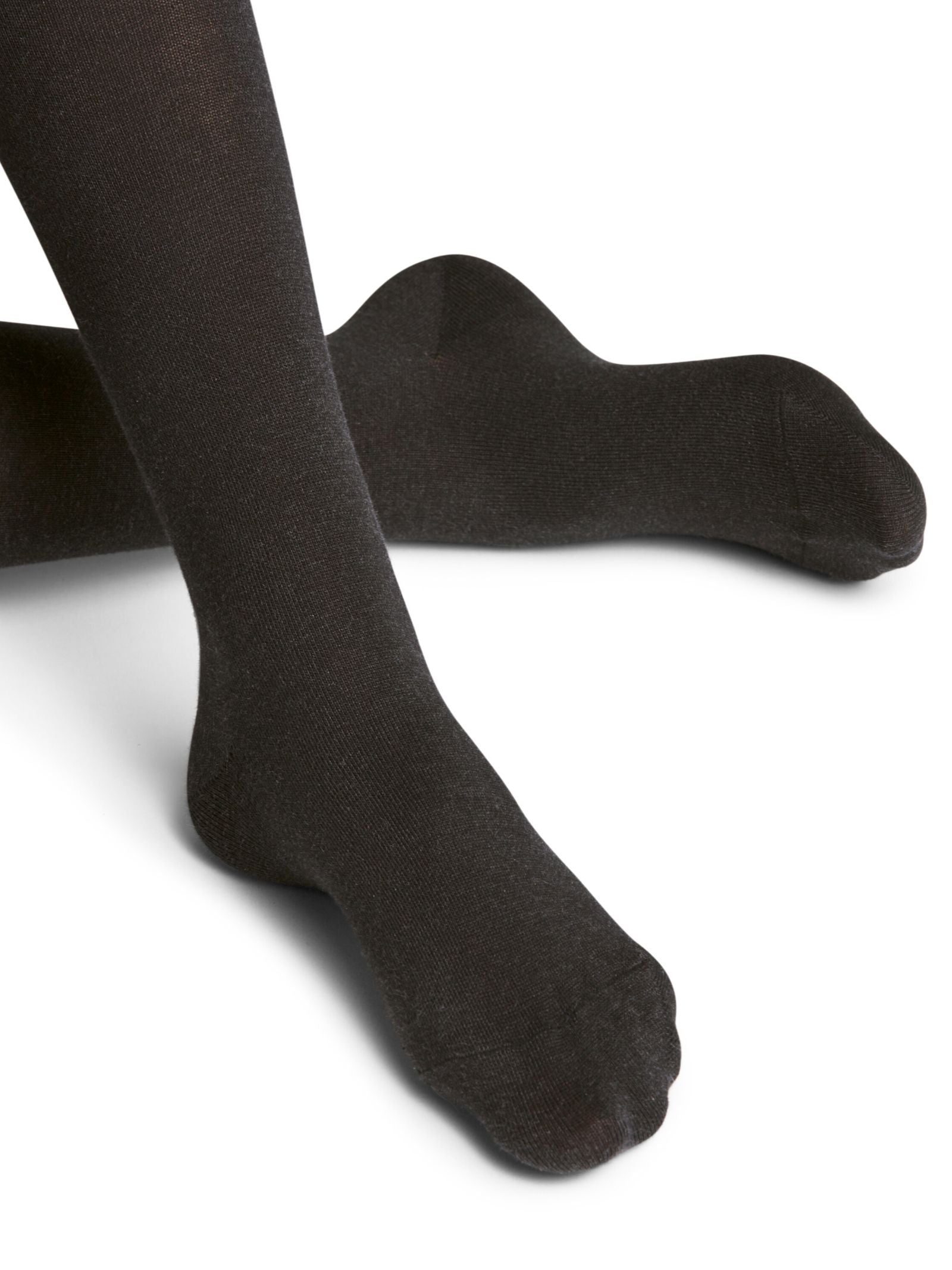 A close-up shows a person wearing FALKE Cotton Tights in anthracite, with crossed lower legs and feet on a white surface; only the sustainably crafted tights are visible.
