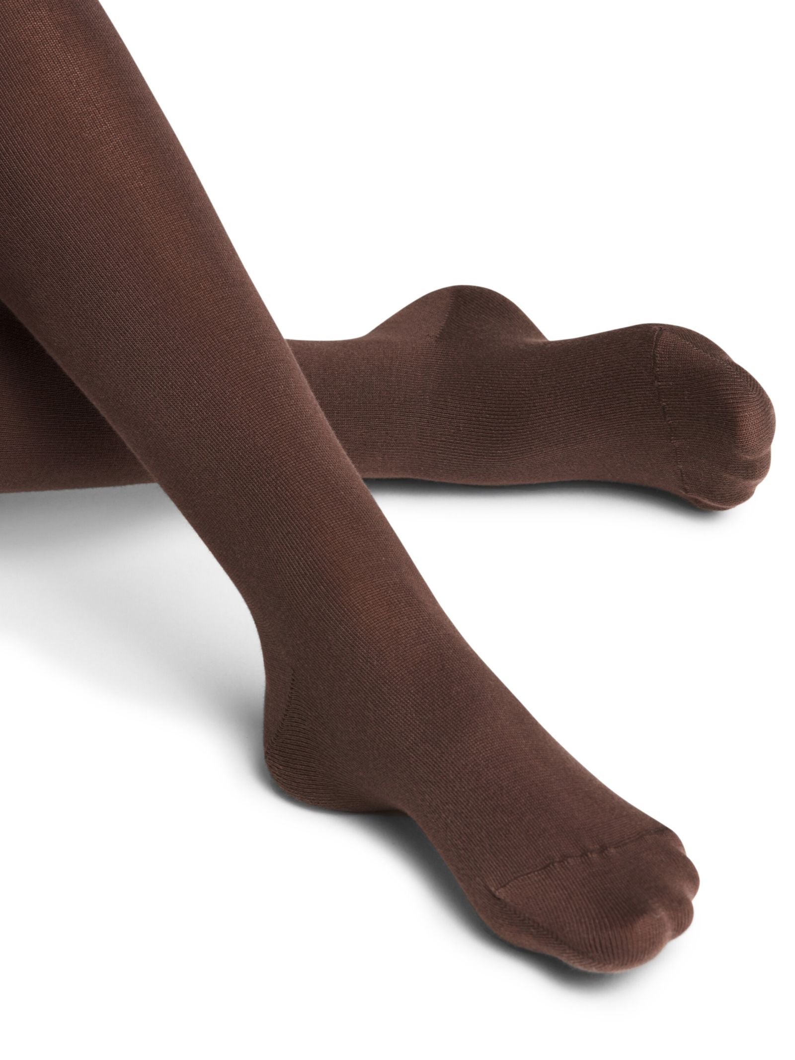 A close-up shows legs in FALKE Cotton Tights in dark brown, feet slightly crossed on a white surface, highlighting the comfort of sustainable cotton.