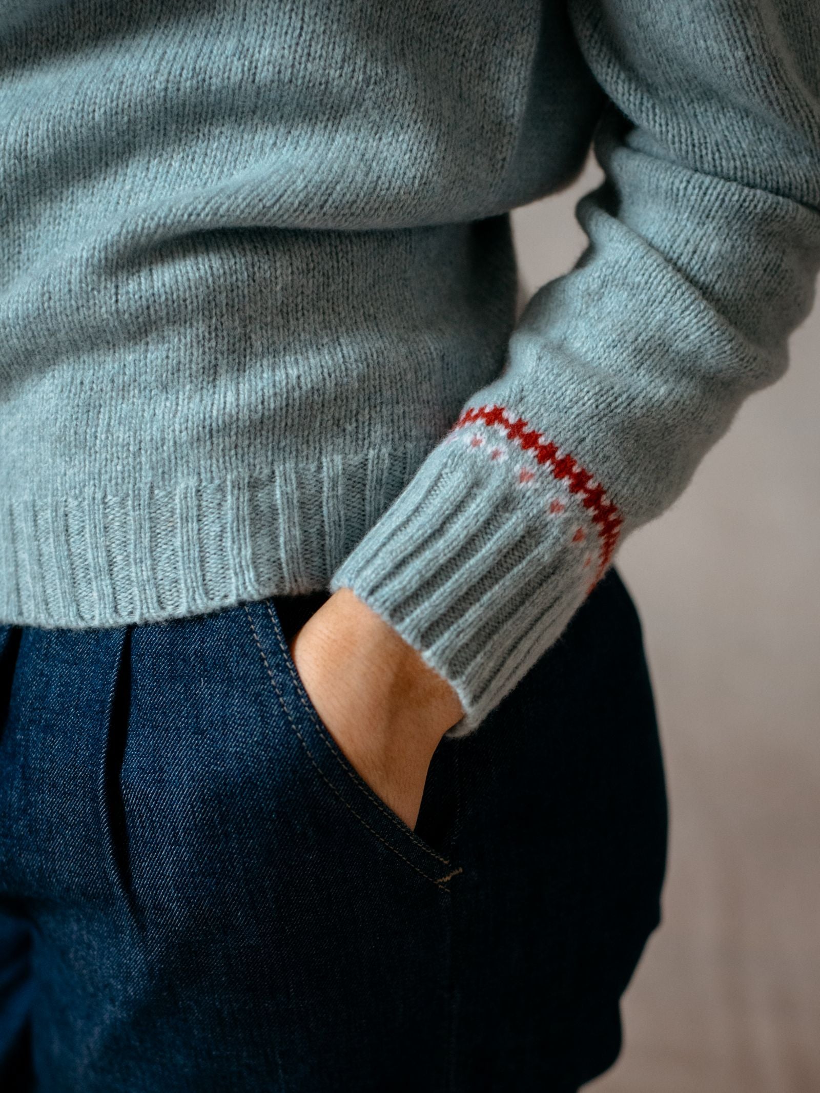A person wears the Campbells of Beauly Geelong Lambswool Strawberry Yoke Jumper with a red and white Fairisle yoke pattern, their hand in the pocket of dark blue jeans.