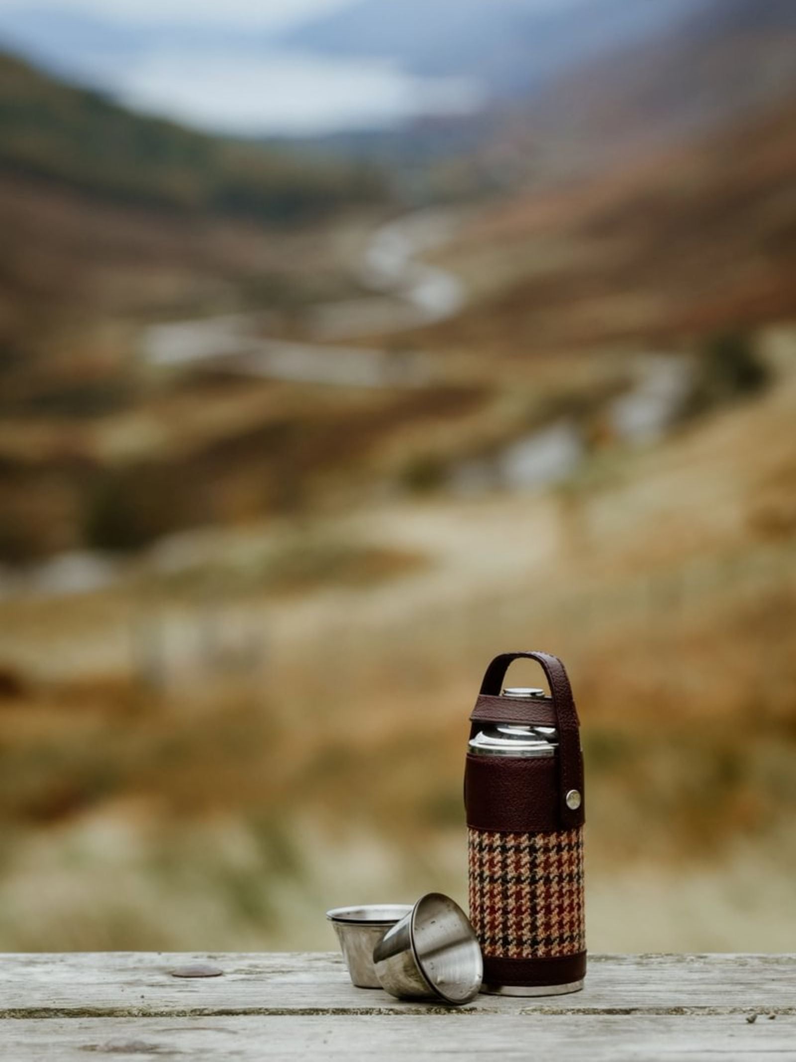 An 8oz Bordeaux Hunter Flask by Campbell's of Beauly x Ettinger, in a plaid case with two metal cups, rests outdoors on wood, highlighting British craftsmanship amid rolling hills and a winding road.
