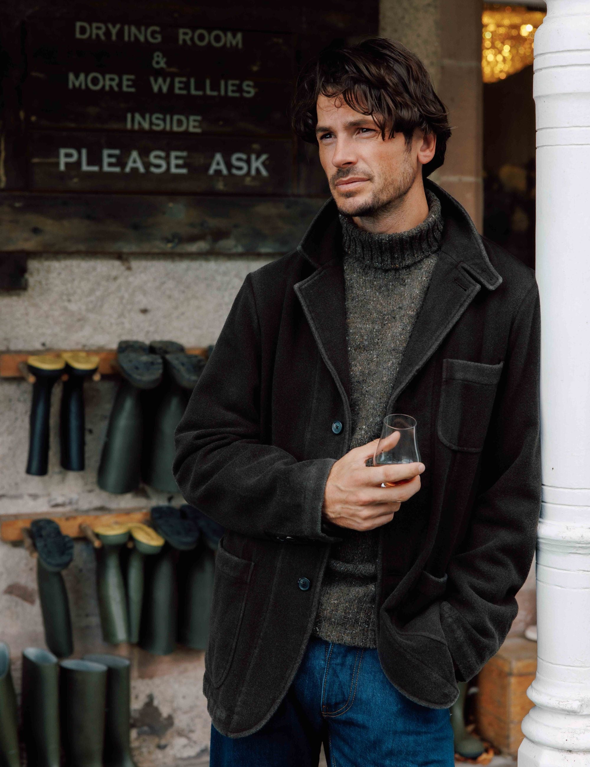 A man with wavy dark hair, wearing a dark jacket and gray sweater, holds a drink and stands in front of a wall with boots and a sign reading “DRYING ROOM & MORE WELLIES INSIDE PLEASE ASK.”.