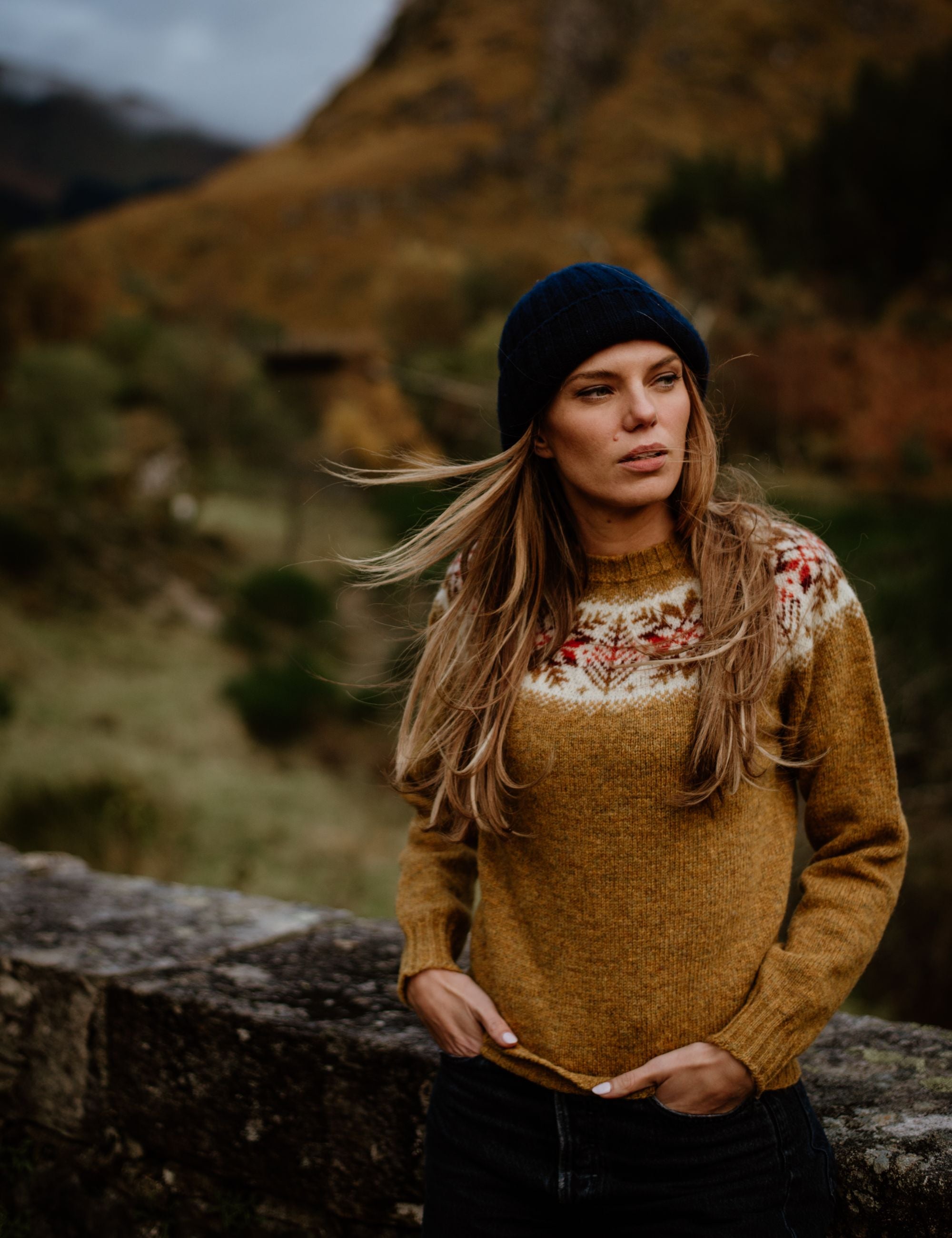 A woman with long blonde hair wears a mustard-colored sweater with a patterned yoke and a navy beanie, standing outdoors by a stone wall with hills and greenery in the background.