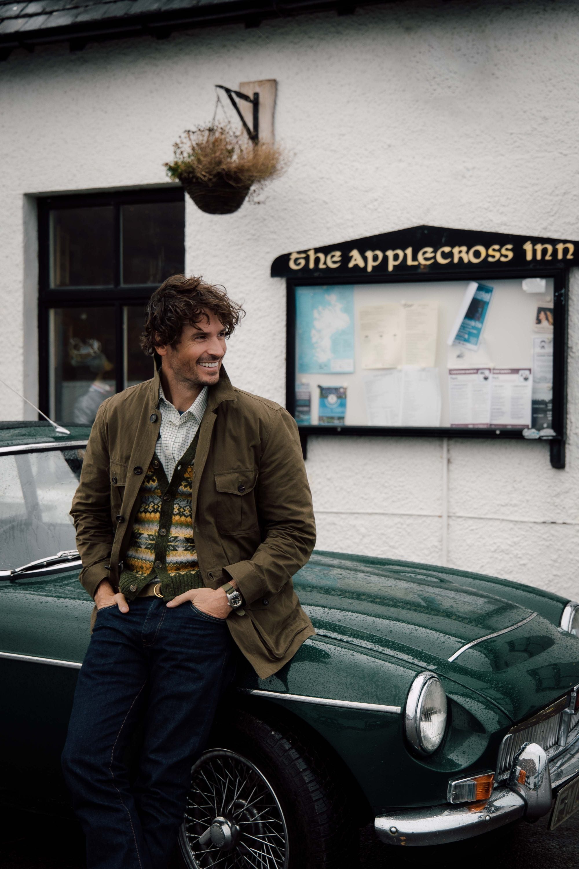 A man wearing a green jacket and patterned sweater smiles while leaning against a vintage green car parked in front of The Applecross Inn, with a notice board and flower pot on the wall behind him.