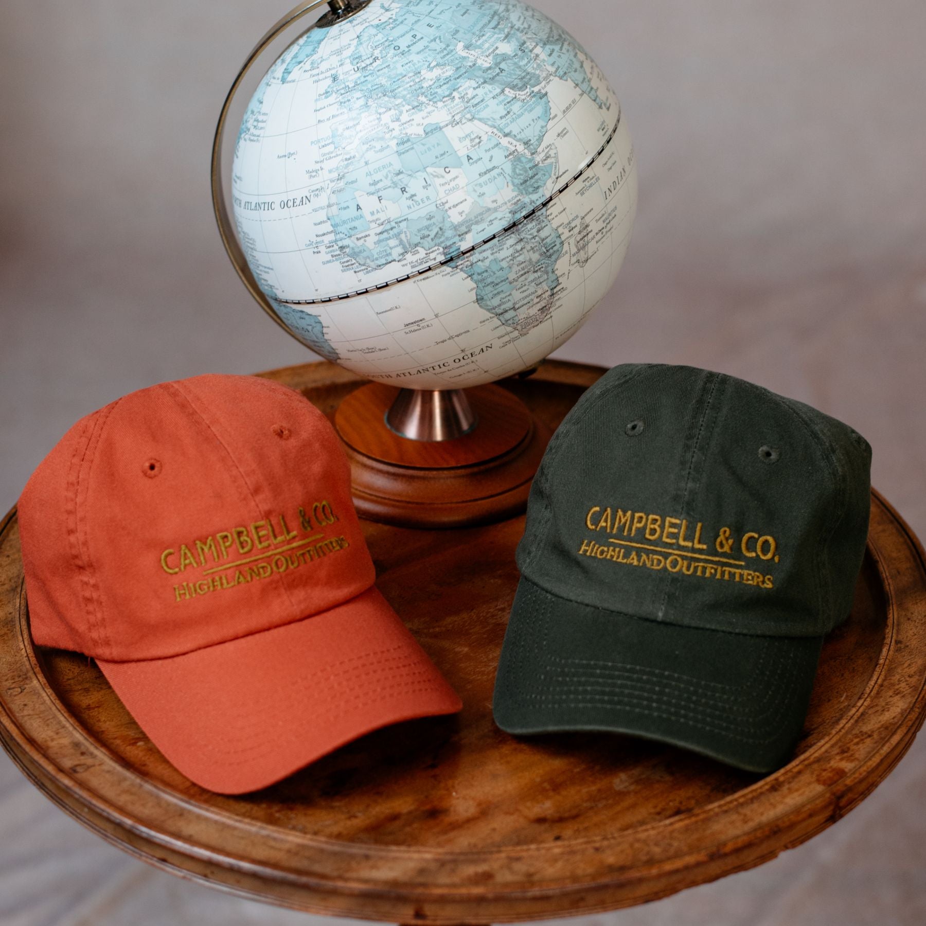 Two embroidered baseball caps, one orange and one dark green, lie on a round wooden table next to a small globe. The caps are labeled CAMPBELL & CO. HIGHLAND OUTFITTERS.