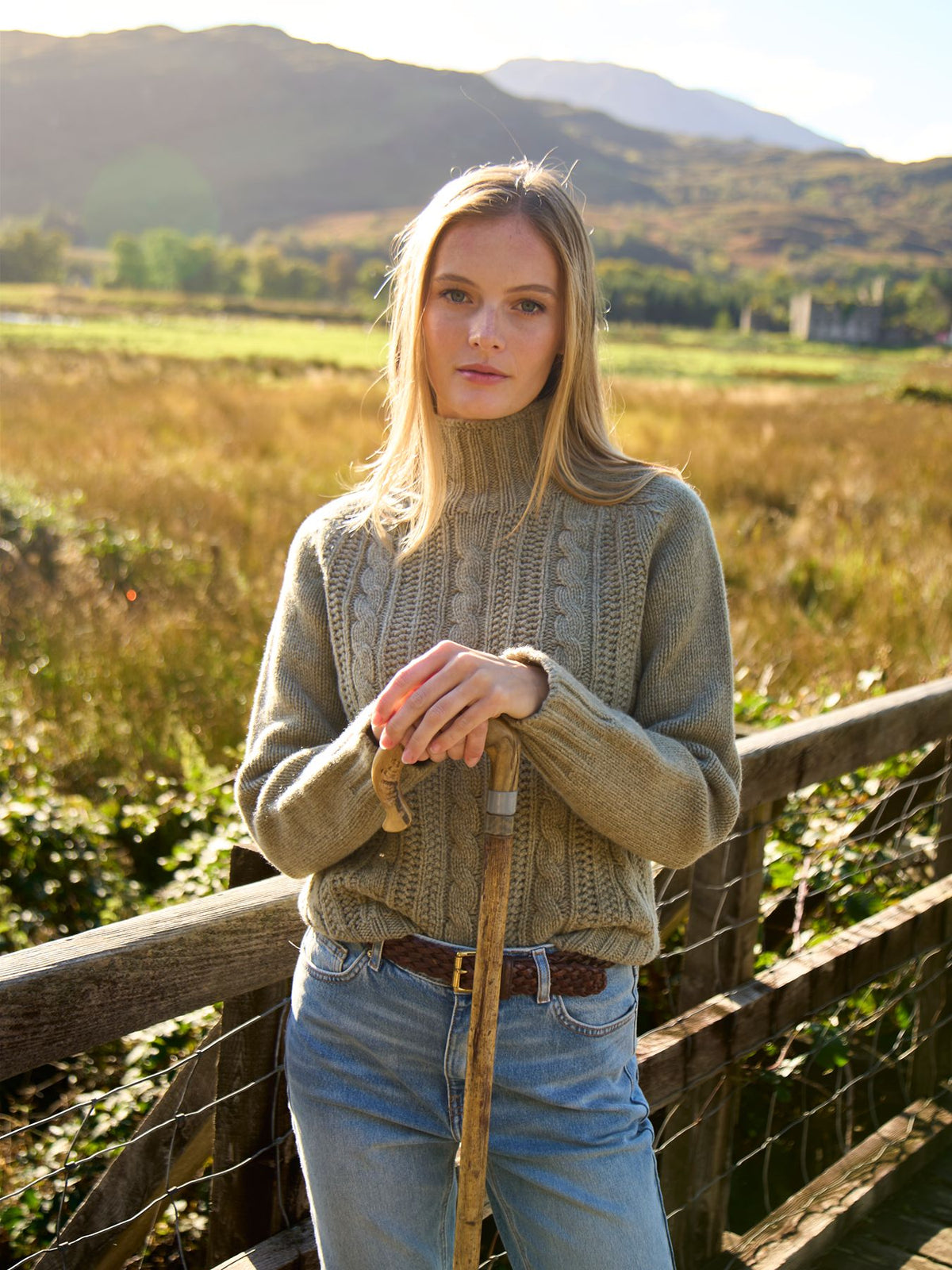 A woman wearing a cable-knit jumper and jeans holds a wooden walking stick whilst leaning on a wooden fence in a field.