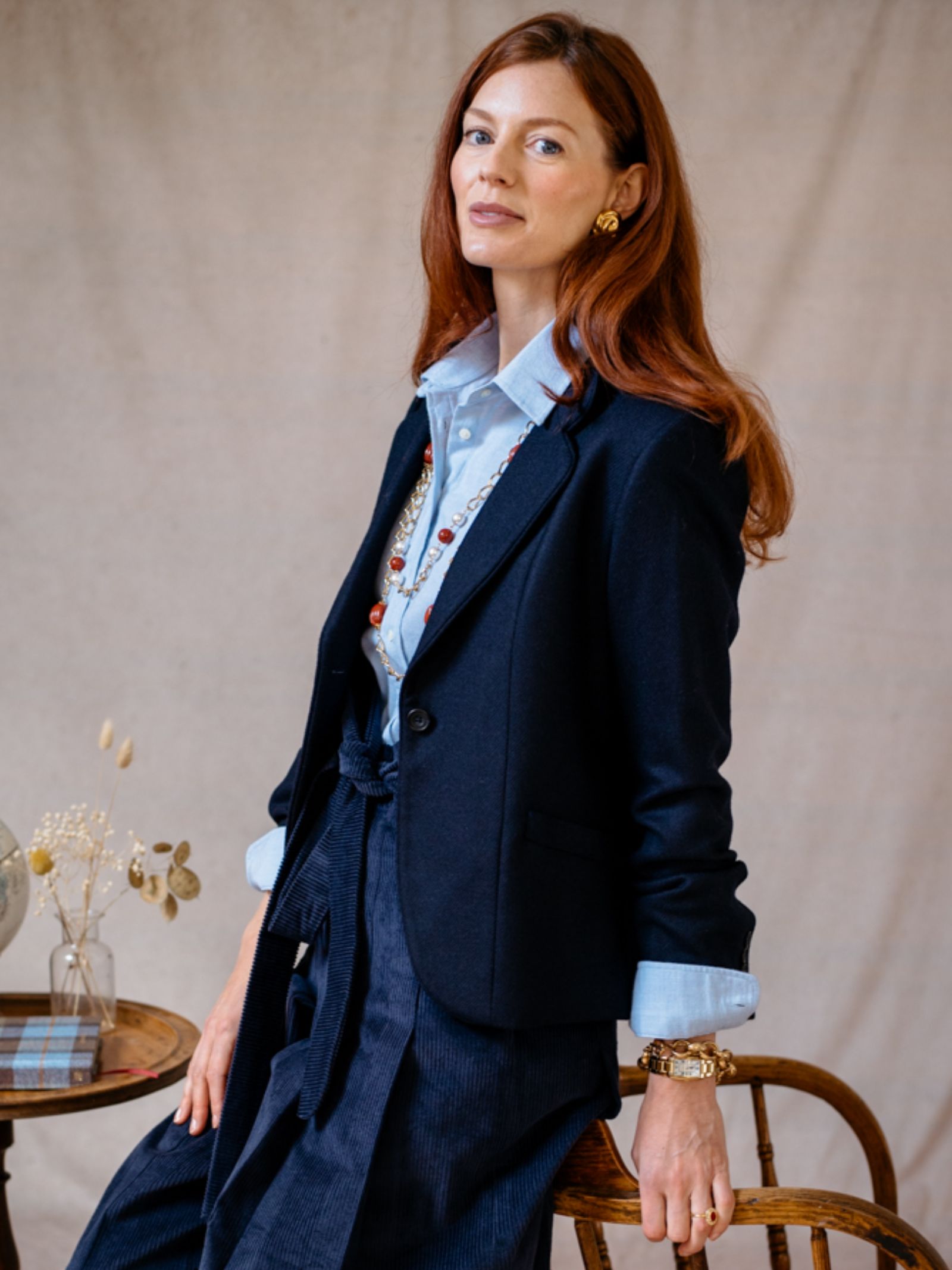 A woman with long red hair wears the Campbells of Beauly House Jacket over a light blue shirt and gold jewelry, seated on a wooden chair beside dried flowers, capturing timeless style against a neutral backdrop.
