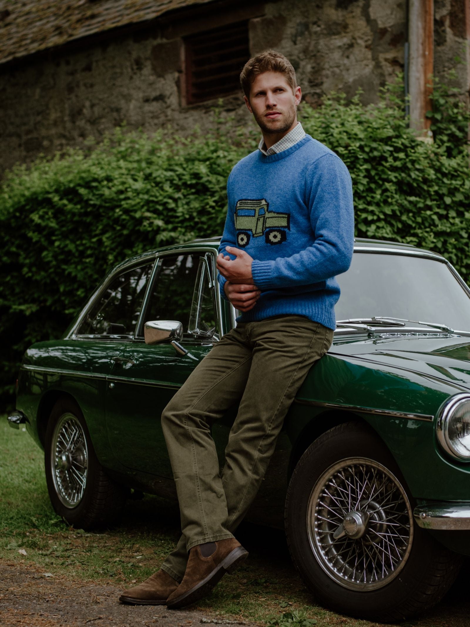 Wearing the Campbells of Beauly Land Rover Defender Jumper, a man leans on a vintage green car near greenery and an old stone building, embodying classic British motoring style.