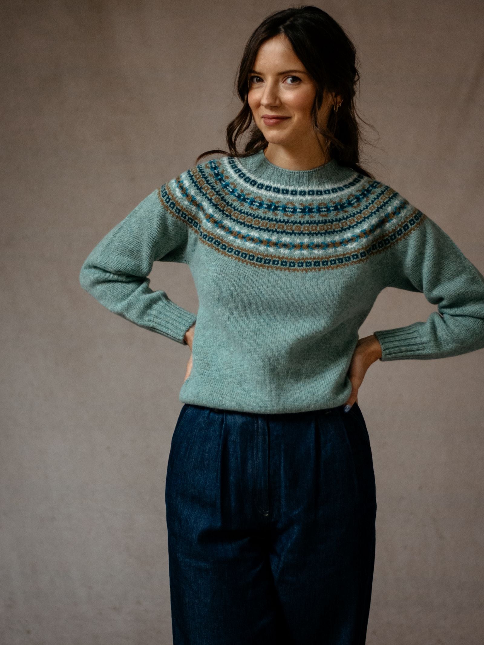 A woman poses with hands on hips, wearing the Campbells of Beauly Fairisle Crew Jumper in light blue superfine Geelong lambswool with a circular pattern, paired with dark blue high-waisted pants, set against a neutral background.