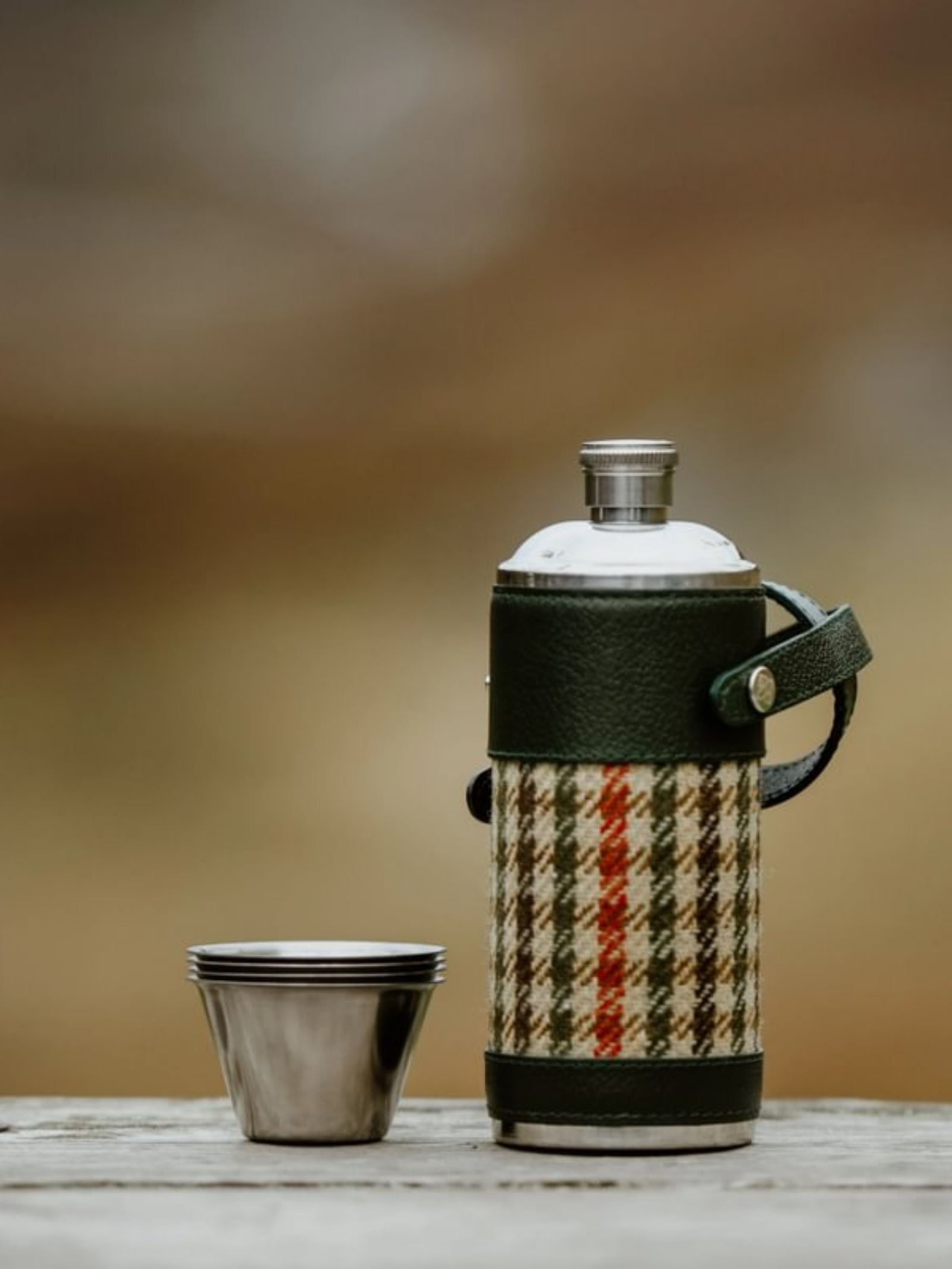 An 8oz Forest Green Hunter Flask by Campbell's of Beauly x Ettinger, with plaid cover and Capra leather accents, rests on wood beside a small metal cup, highlighting British craftsmanship against a softly blurred outdoor backdrop.