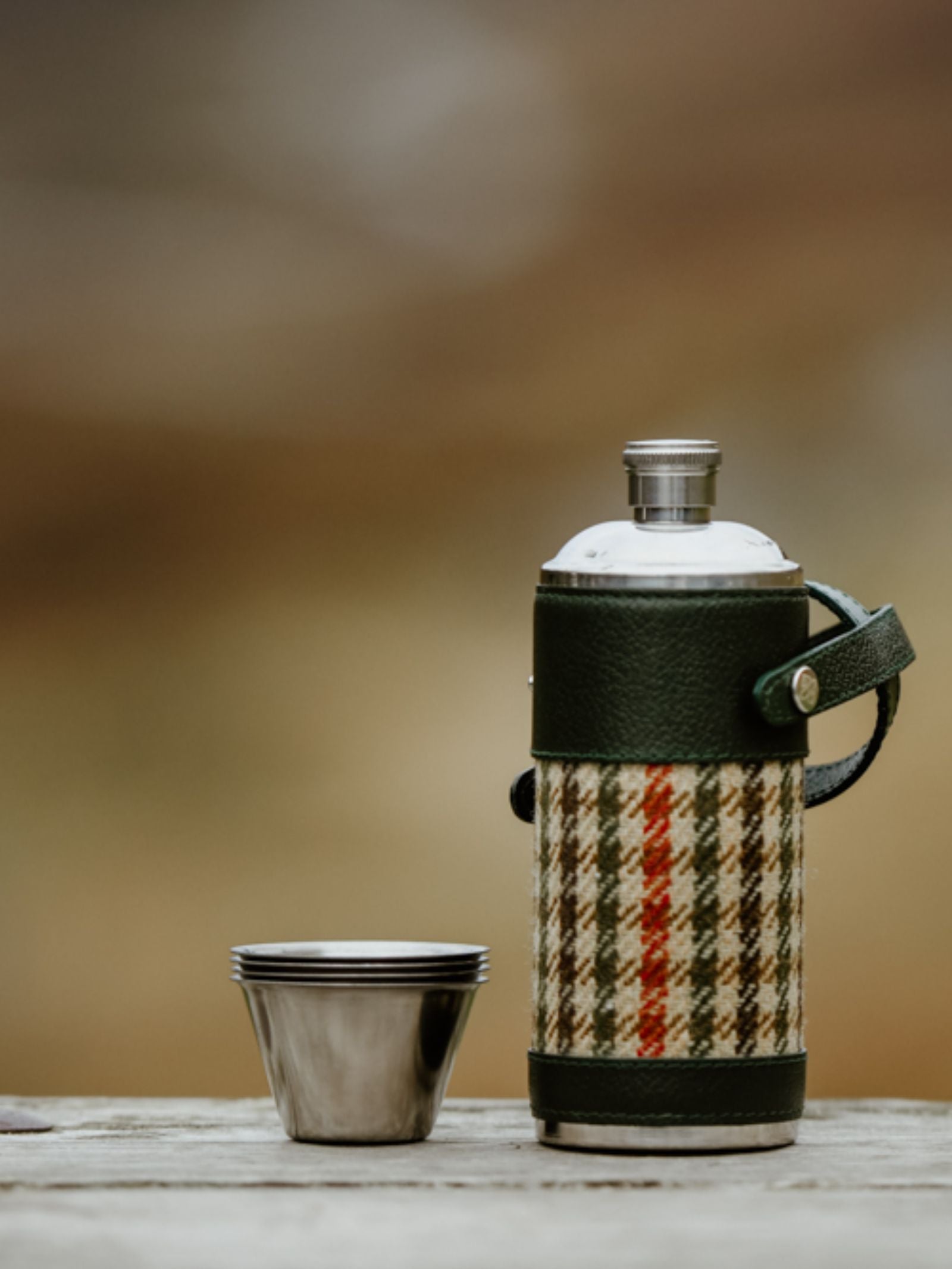 The 8oz Bordeaux Hunter Flask by Campbell's of Beauly x Ettinger, wrapped in Capra goat leather and plaid fabric, rests on wood beside two stacked stainless steel cups, set against a softly blurred outdoor backdrop.
