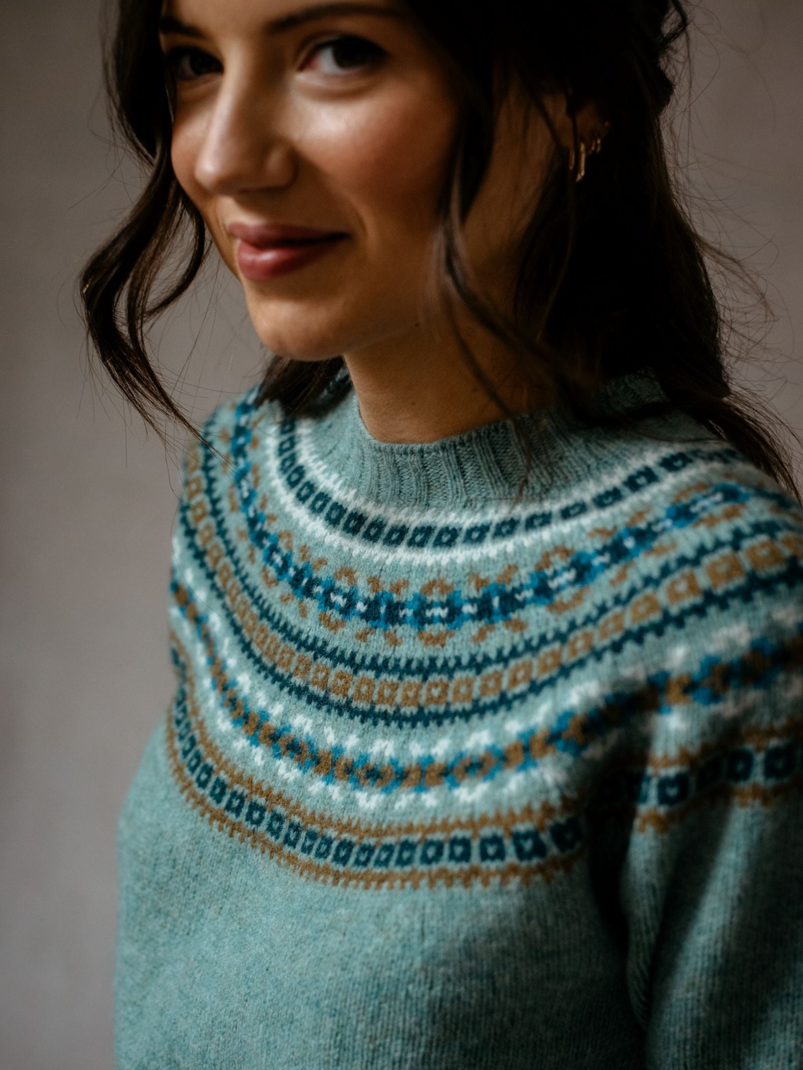 A woman with wavy brown hair smiles gently, facing slightly to the side, wearing a light blue Campbells of Beauly Fairisle Crew Jumper with an intricate, colorful pattern around the yoke.