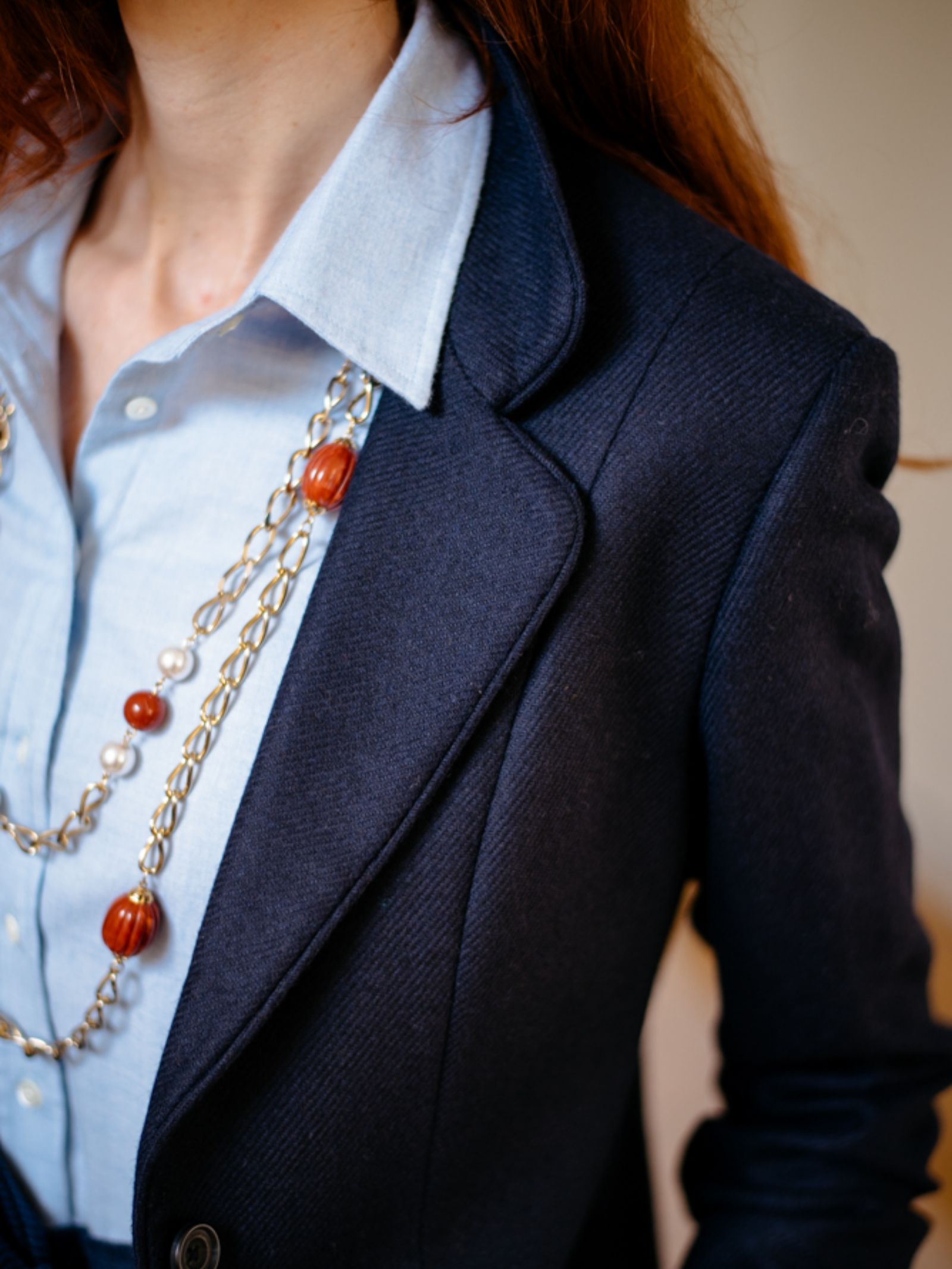 A person wears a Brushed Cotton Shirt in light blue by Campbells of Beauly, styled with a navy blazer and a long gold chain necklace with red and pearl-like beads. Only the upper torso and part of long brown hair are visible—a timeless look.