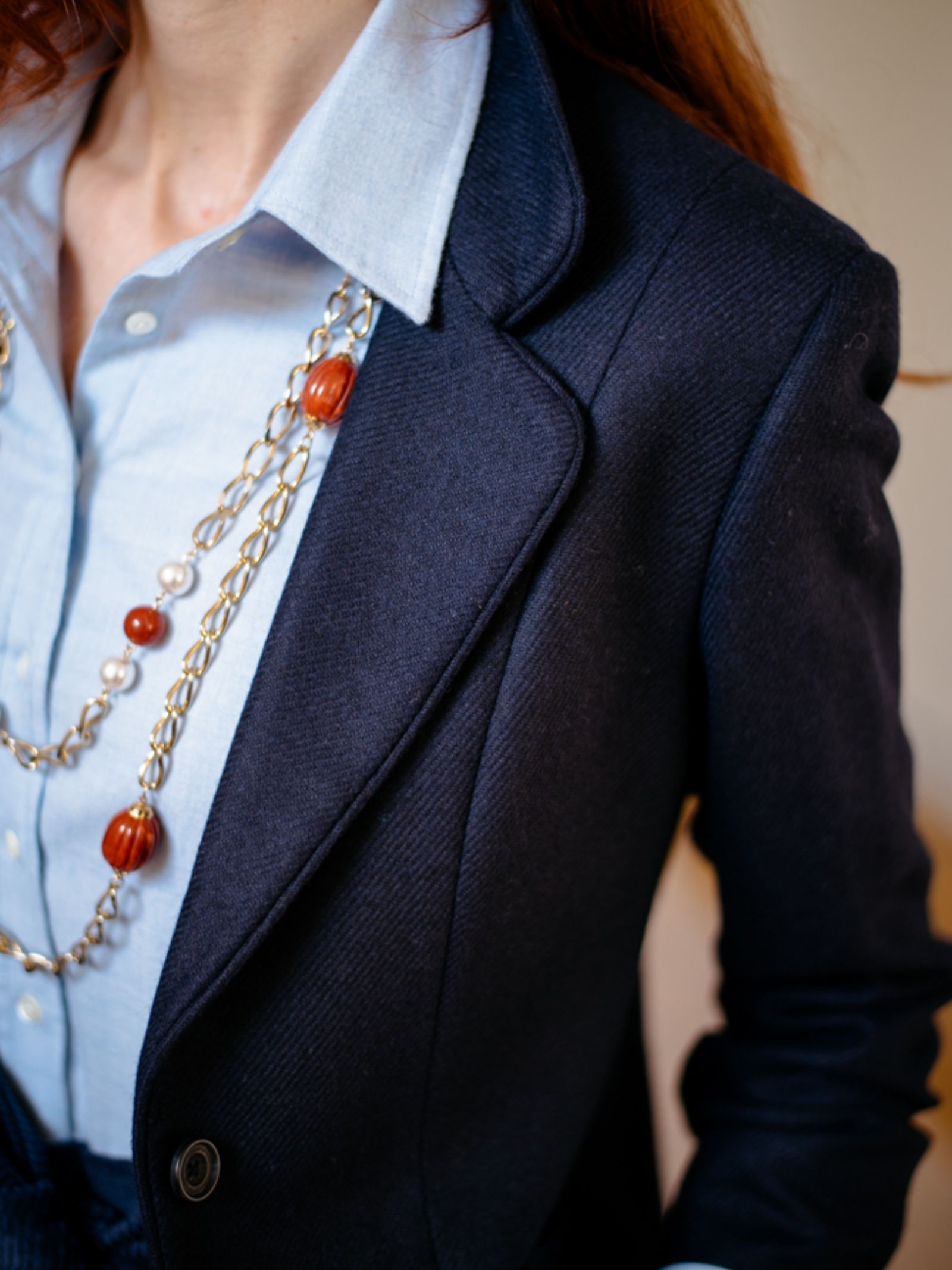 A person wearing the House Jacket by Campbells of Beauly—a navy herringbone tweed blazer over a light blue shirt and finished with a long gold chain necklace with brown beads—exudes timeless style.