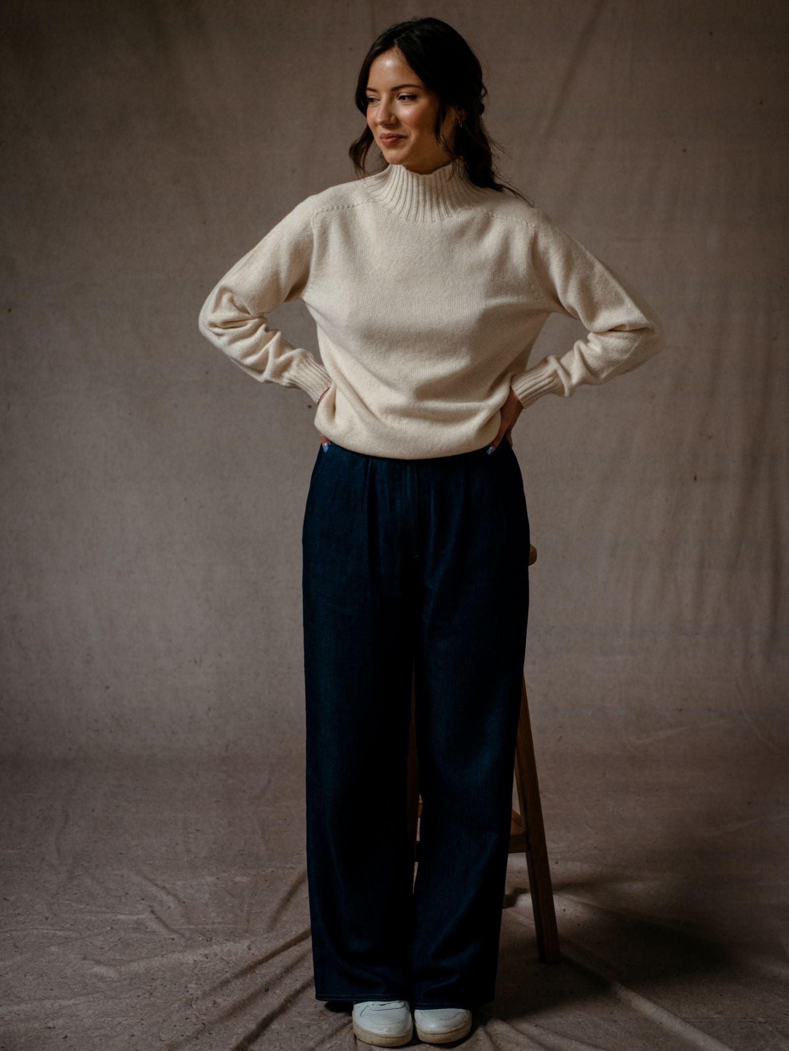 A woman smiles and poses in Campbells of Beauly Wide Leg Jeans, a cream sweater, and white shoes against a plain neutral backdrop.