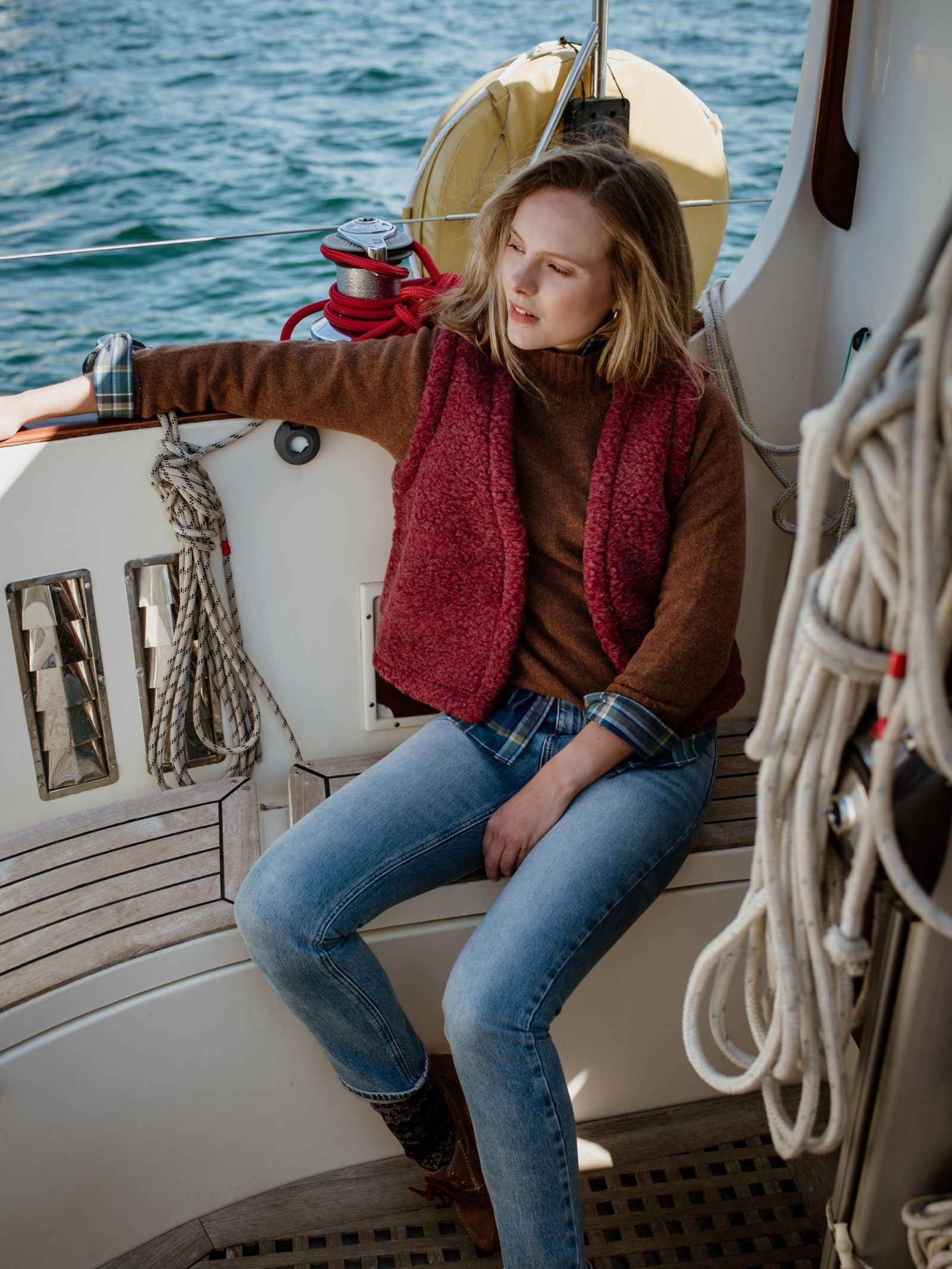 A woman in a Campbells of Beauly Wool Shrug, red vest, and blue jeans sits relaxed on a boat with coiled ropes around her and the ocean in the background.
