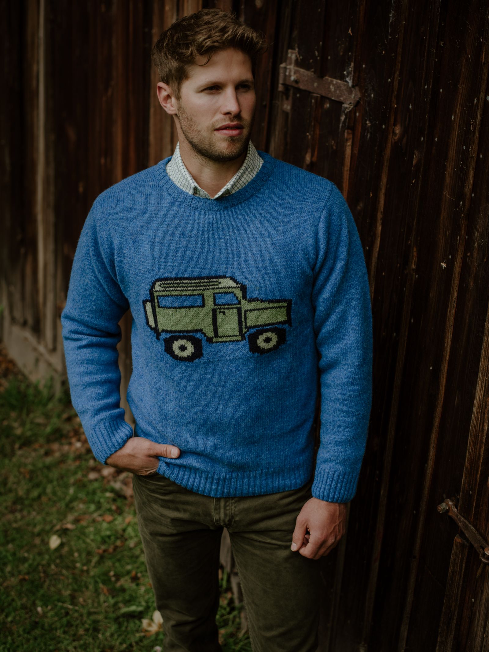 A man stands outside by a wooden wall, wearing Campbells of Beaulys Land Rover Defender Jumper—a blue sweater featuring a classic Defender graphic—paired with olive green pants, embodying timeless British motoring style.