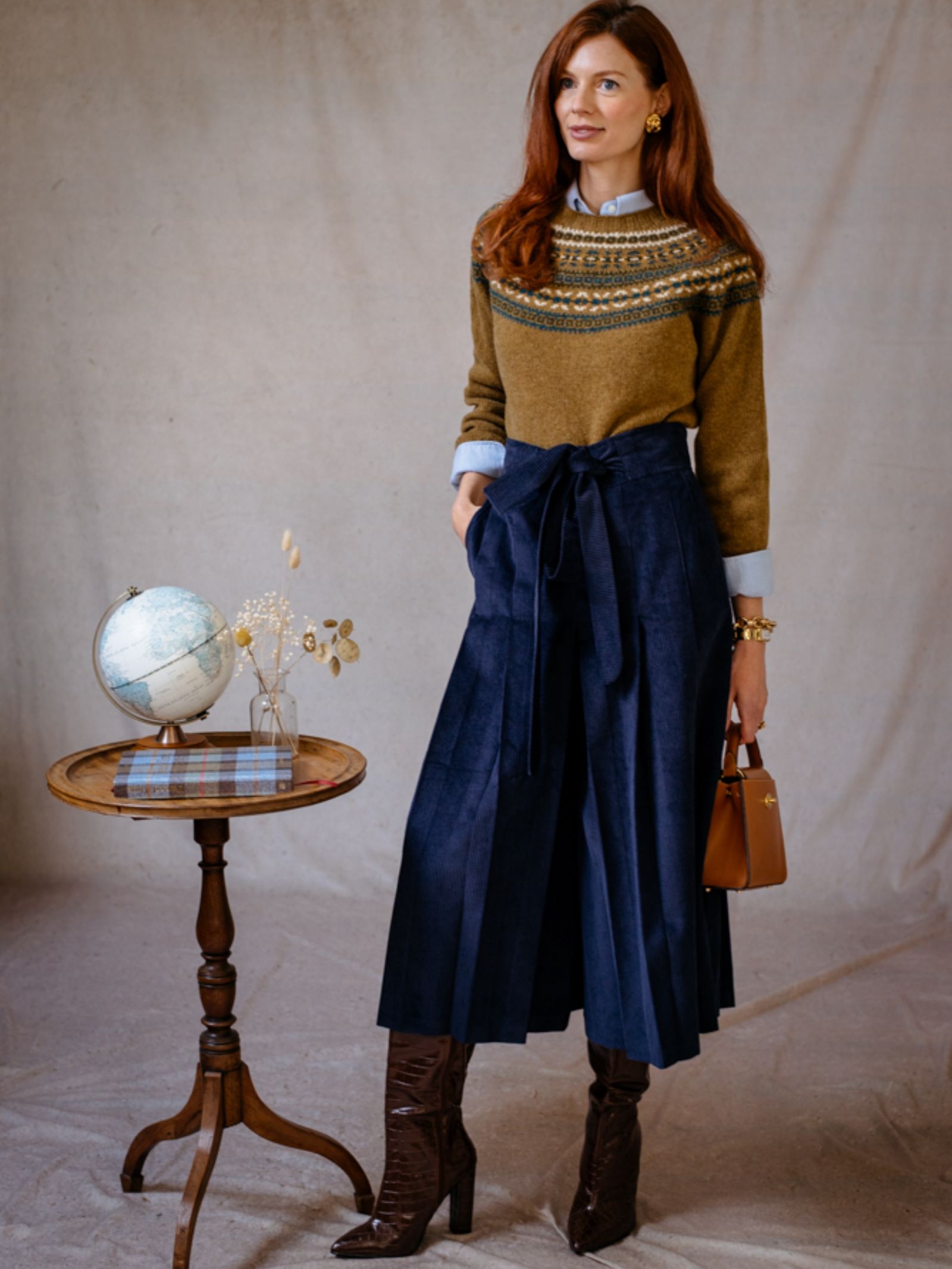 A woman with long red hair wears the Campbells of Beauly Fairisle Crew Jumper over a collared shirt, navy culottes, and brown boots. She stands by a table with a globe, dried flowers, and books, holding a brown handbag.