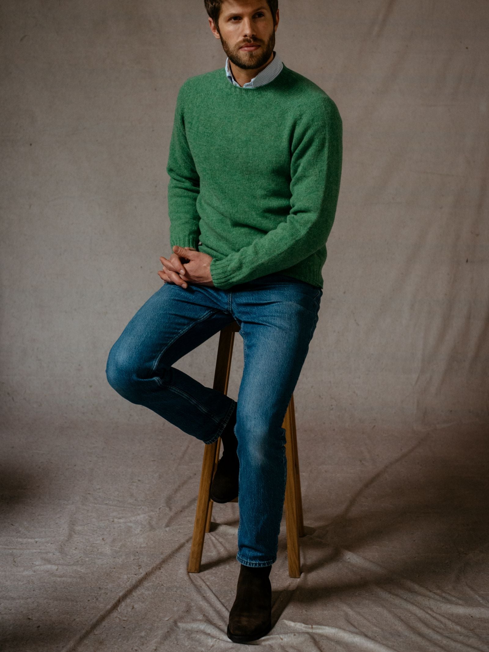A man in a green Campbells of Beauly Shetland Jumper made from soft lambswool, blue jeans, and dark boots sits on a wooden stool against a neutral background, gazing slightly to his left.
