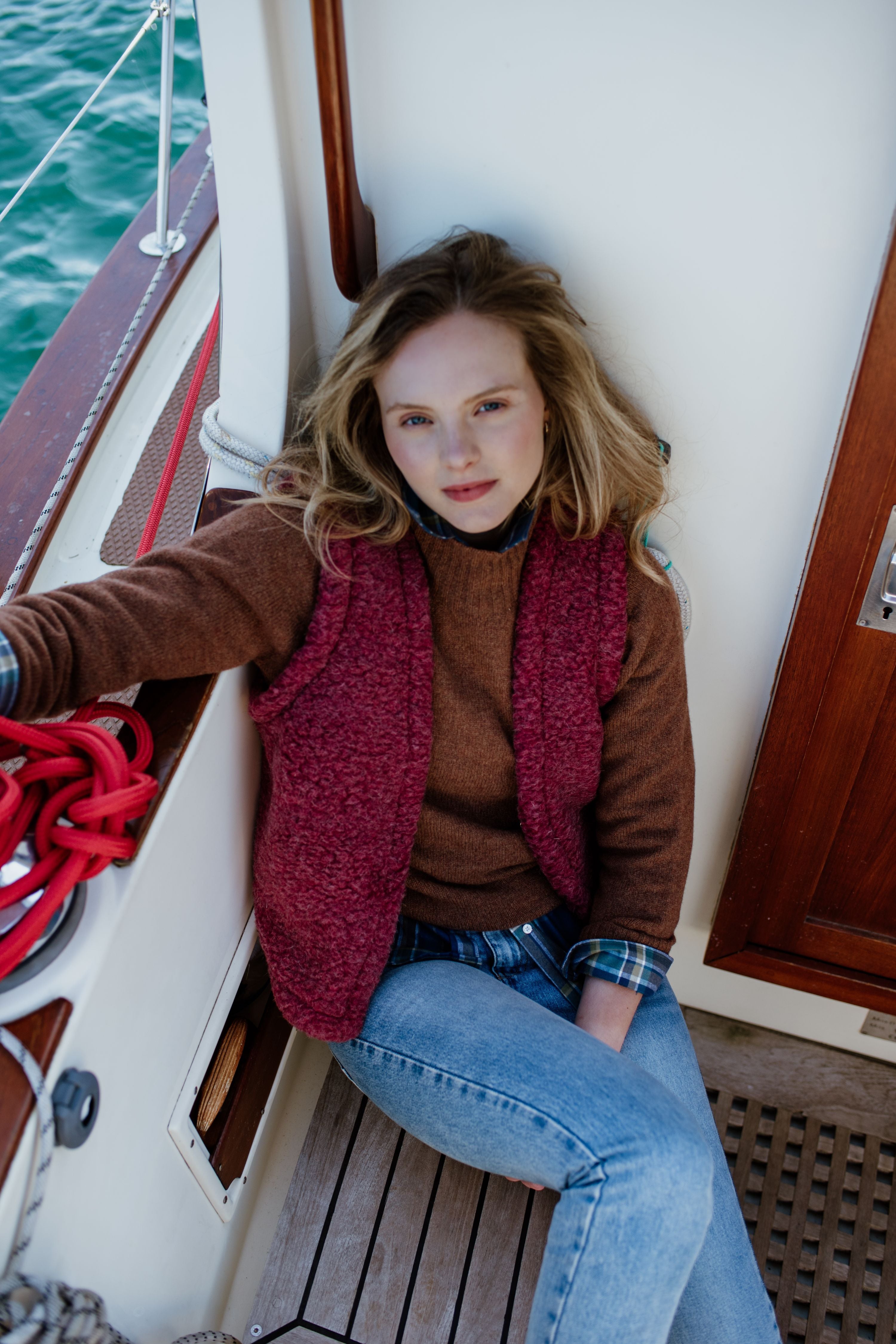 A young woman with long blonde hair, wearing a brown sweater, red vest, and jeans, sits relaxed on the wooden deck of a boat near a door, with coiled red rope beside her and water visible in the background.