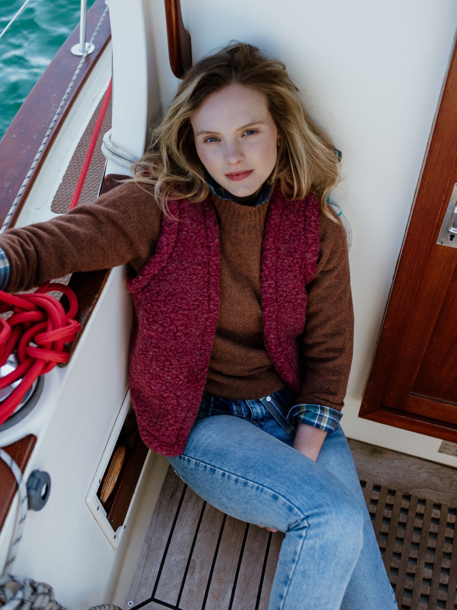 A young woman with blond hair, wearing the Campbells of Beauly Wool Shrug for lightweight warmth and a red vest with jeans, sits relaxed on a boat floor by a wooden door, gazing at the camera.