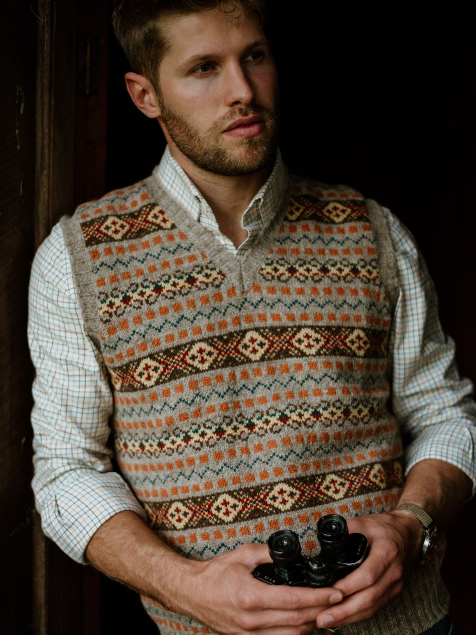 A man with short brown hair and a trimmed beard wears a patterned wool sweater vest over a Campbells of Beauly Windowpane Shirt. Holding black binoculars, he looks pensively to the side near a wooden doorway against a dark background.