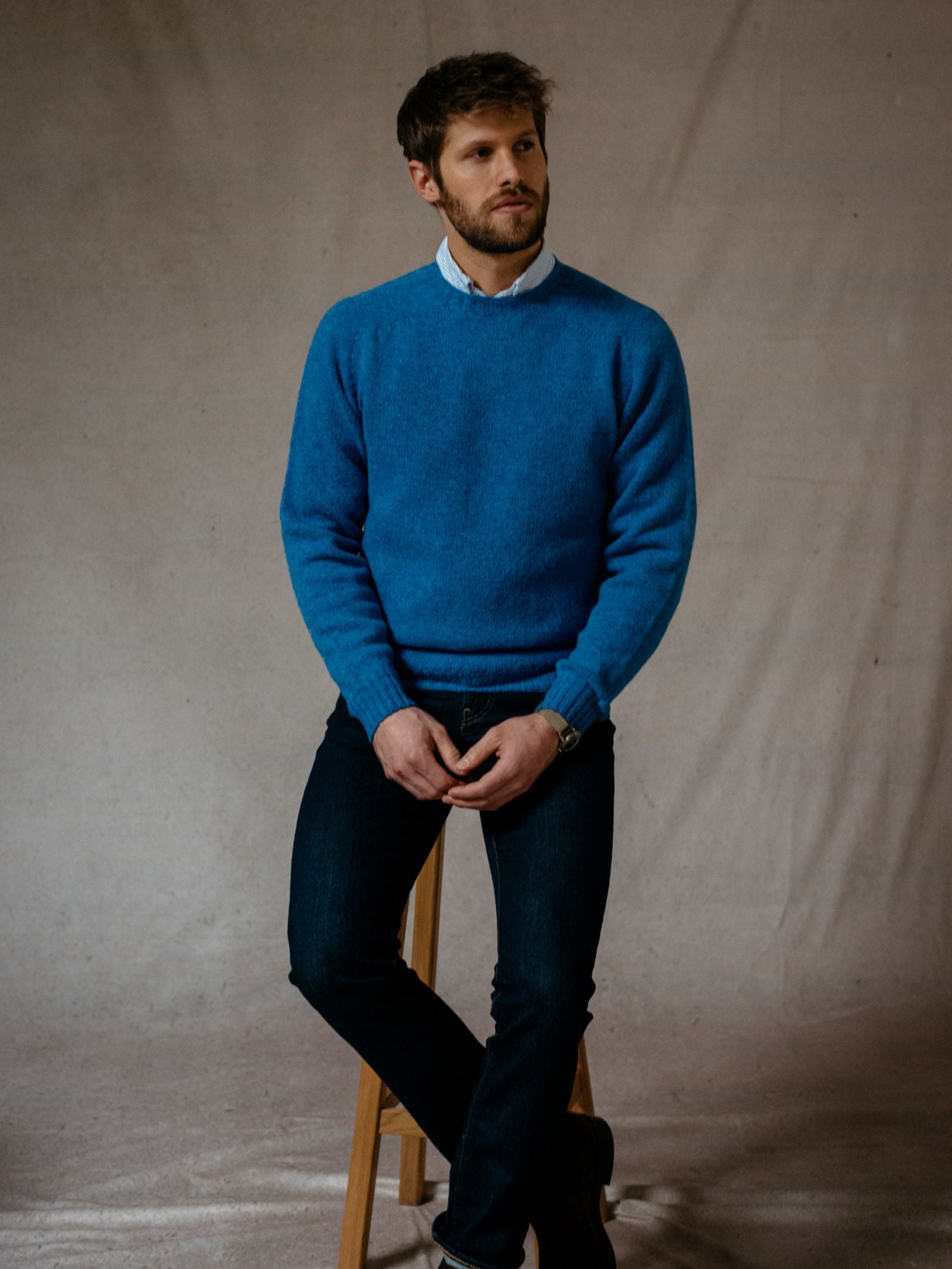A man with short brown hair and a beard sits on a wooden stool, wearing a bright blue Campbells of Beauly Shetland Jumper over a collared shirt and dark jeans, his hands resting on his lap against a plain neutral background.