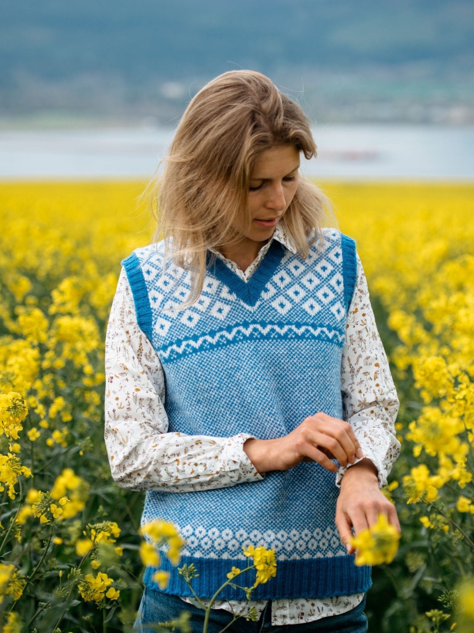 A blonde woman stands in a field of yellow flowers, wearing the Campbells of Beauly Nordic V Tank over a floral shirt. She adjusts her watch while a softly blurred lake and mountains appear in the background.