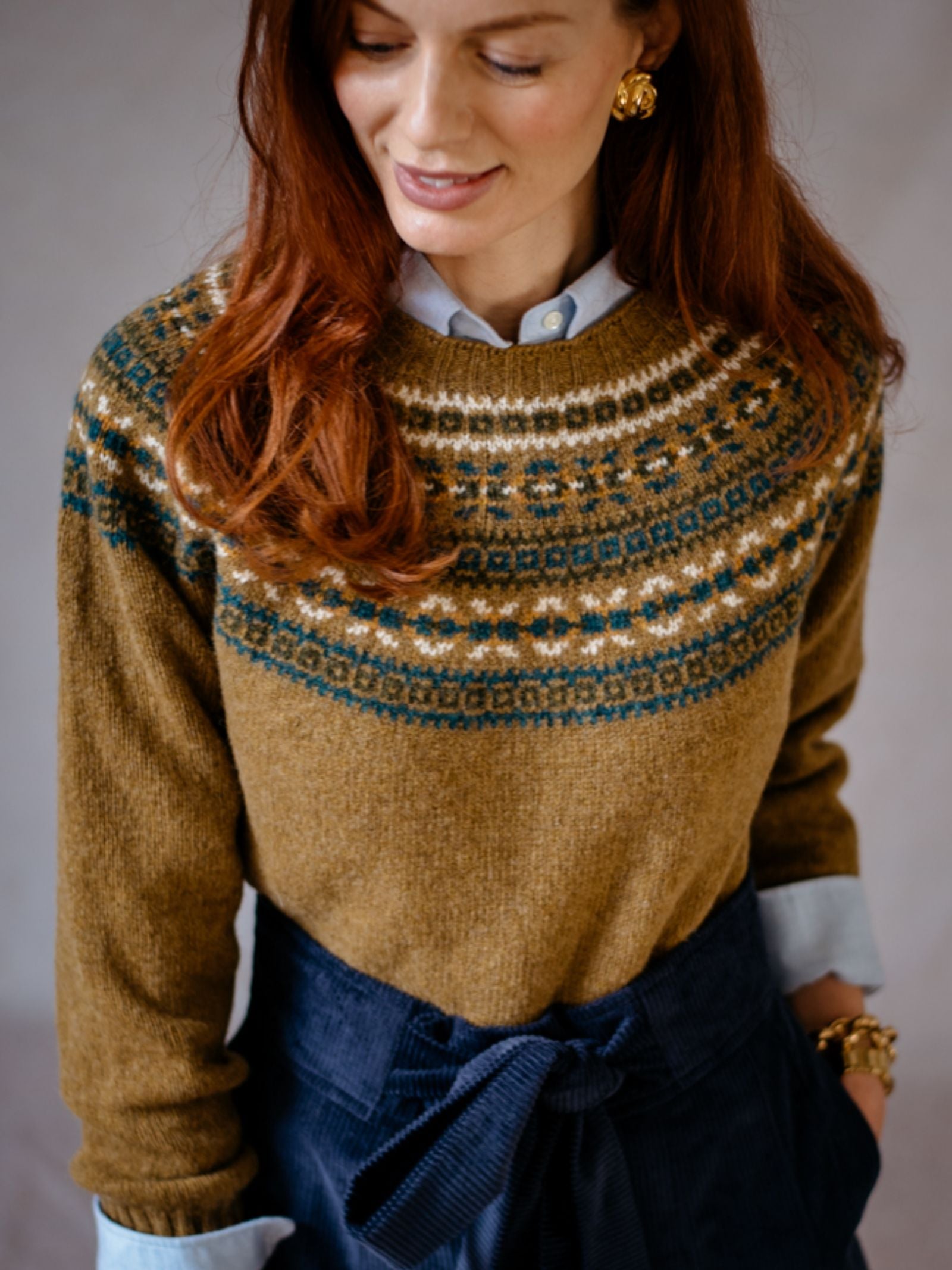 A woman with long auburn hair wears the Campbells of Beauly Fairisle Crew Jumper in a traditional pattern over a light blue shirt and navy high-waisted pants, looking down with a gentle smile.
