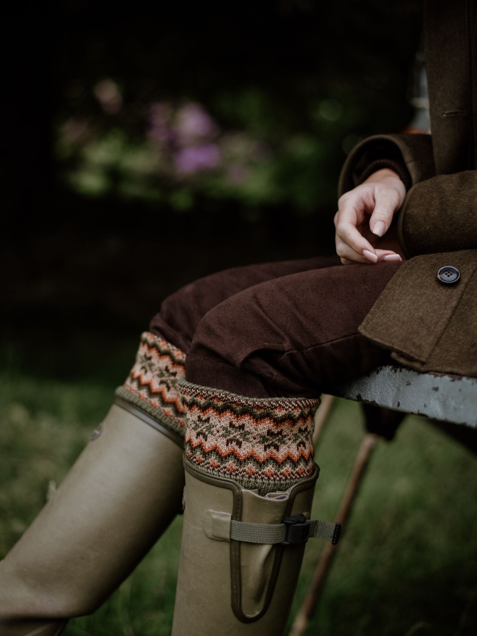 A person outdoors wears Campbells of Beauly Merino Fairisle Shooting Socks peeking from olive green boots and brown pants, with only their lower body and one hand visible amid grass and dark foliage.