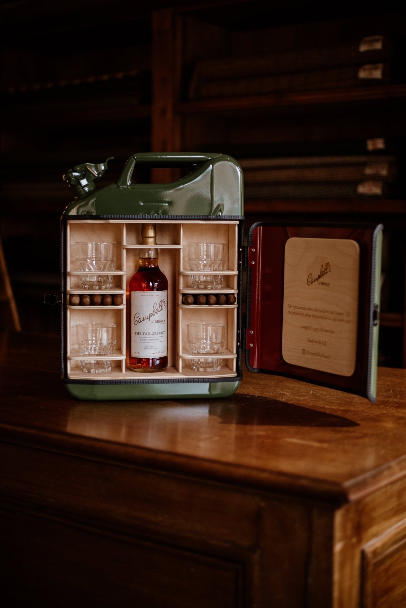 The Connoisseur Upcycled Mini Bar by Campbells of Beauly is a green, handmade UK jerry can repurposed as a portable bar with six glasses, a bottle of Campbells whisky, and branded text on the inner door—all displayed on wood.