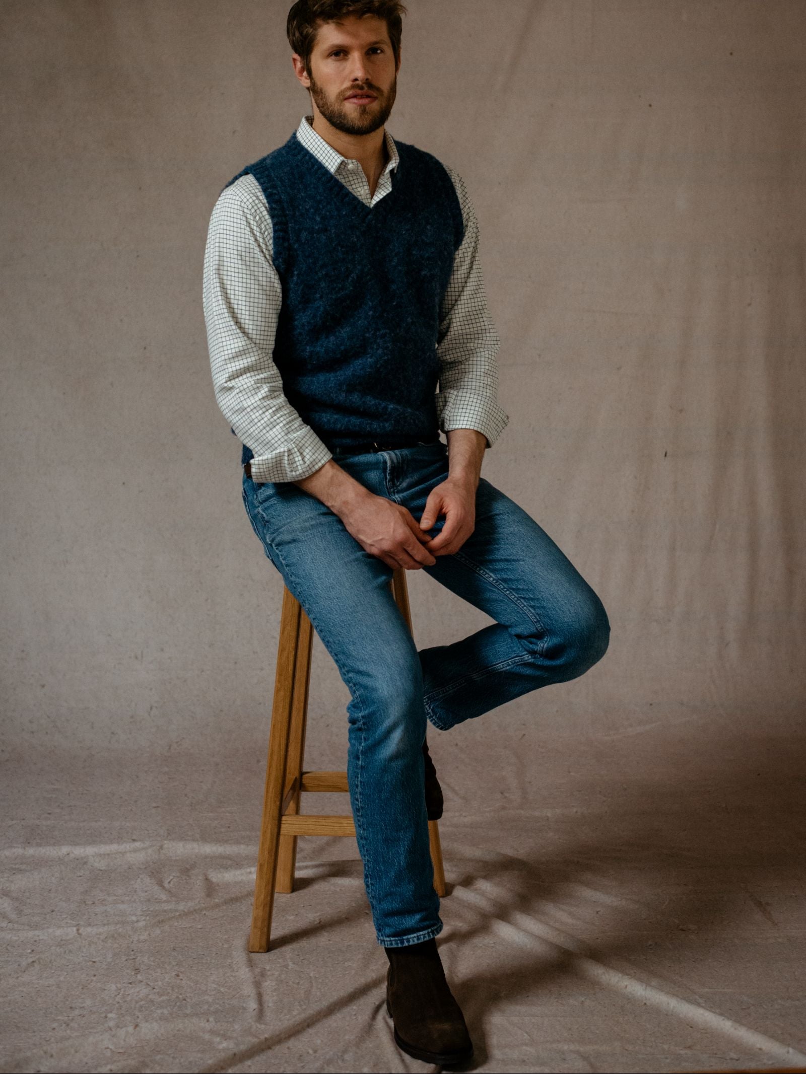 A man with short hair and a beard sits on a wooden stool against a plain background, wearing blue jeans, brown boots, a checked shirt, and the Campbells of Beauly Shetland Slipover, looking directly at the camera.