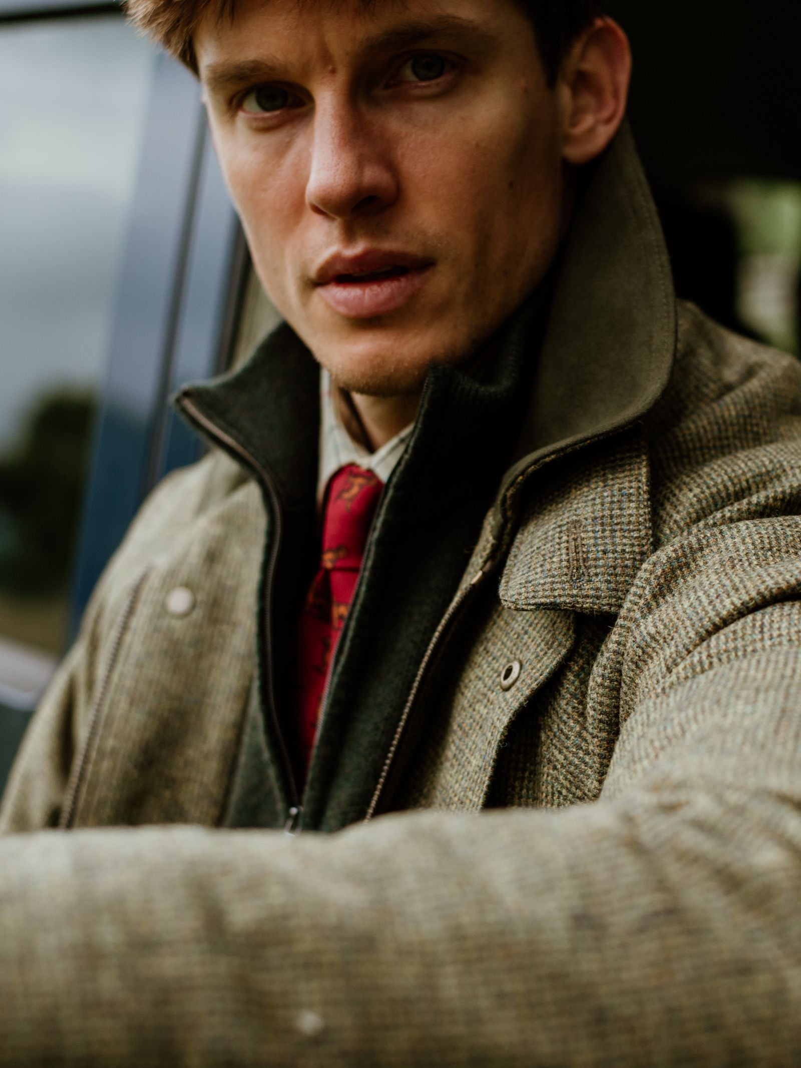 A man with light brown hair, wearing a tweed jacket, Campbell's of Beauly Merino & Cashmere Quarter Zip Jumper, collared shirt, and red tie, sits in a car looking directly at the camera; the background is slightly blurred.