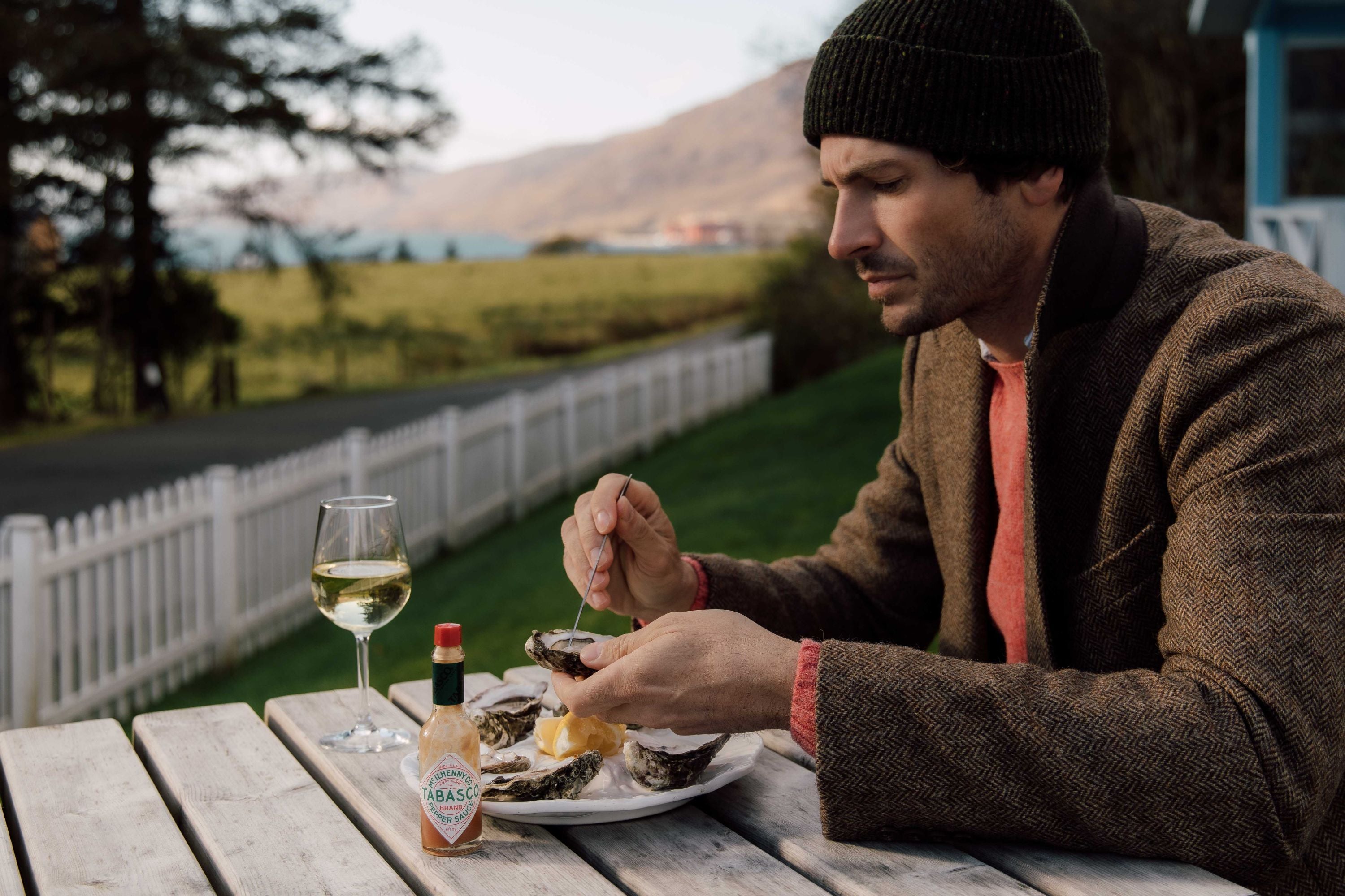 A man wearing a brown coat and black beanie sits at an outdoor table, preparing to eat oysters with lemon. A glass of white wine and a bottle of Tabasco sauce are on the table. Green fields and mountains are visible in the background.