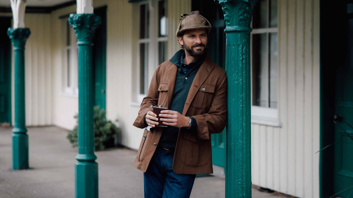 A man wearing a deerstalker hat and brown jacket leans against a green pillar whilst holding a metal flask.