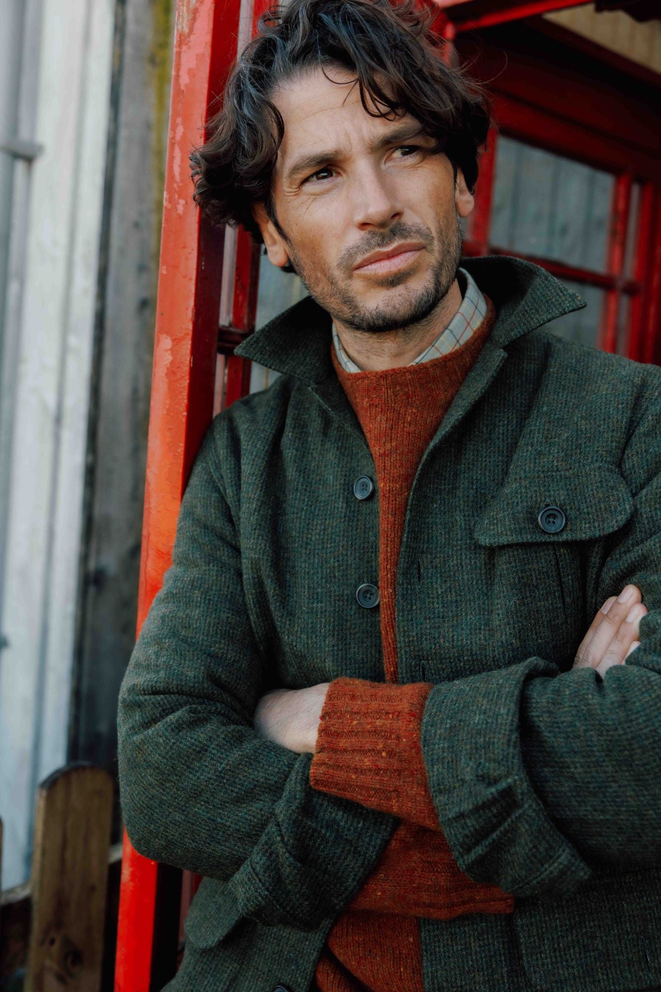 A man with wavy dark hair and stubble stands with arms crossed, wearing a green jacket over an orange sweater and plaid shirt. He leans against a red structure, looking to the side with a thoughtful expression.